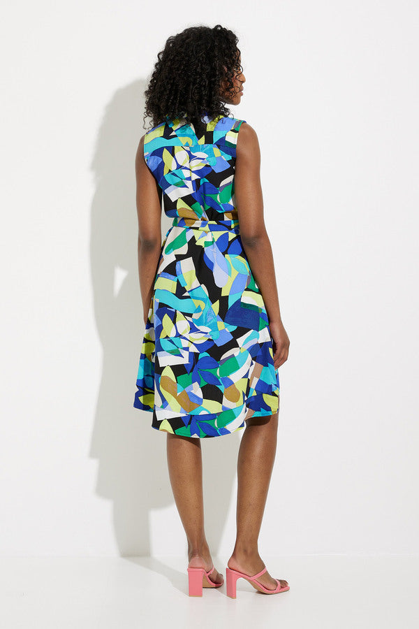 A person with curly hair stands against a white background, wearing the Joseph Ribkoff Button Front Dress Style 232090. This sleeveless dress boasts a bold geometric print perfect for any tropical getaway and is paired with pink open-toe heels. The dress features a collar and buttons down the front, as the individual stands with one hand resting by their side.