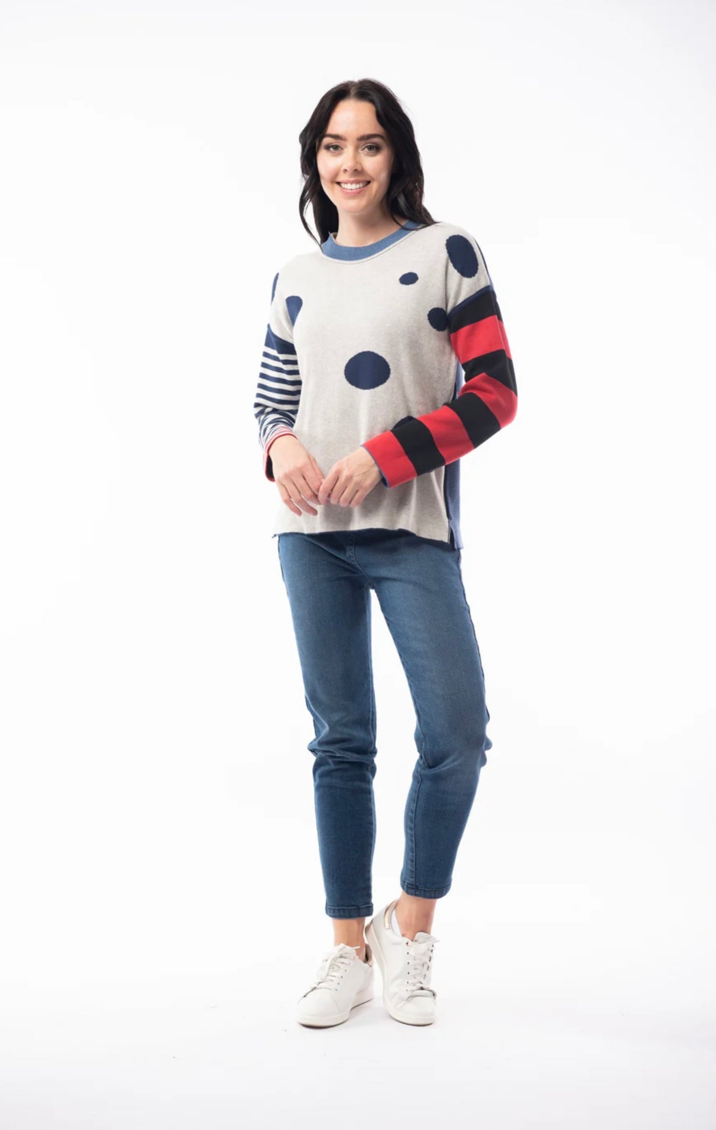 A woman with long dark hair is wearing the ORIENTIQUE - REVERSIBLE ROUND NECK SPOT/STRIPE JUMPER (1229) made of organic cotton, featuring a navy blue background adorned with white polka dots and red striped sleeves. She's paired it with blue jeans and white sneakers, and stands in a casual pose, slightly leaning back with her hands behind her. The background is plain white.