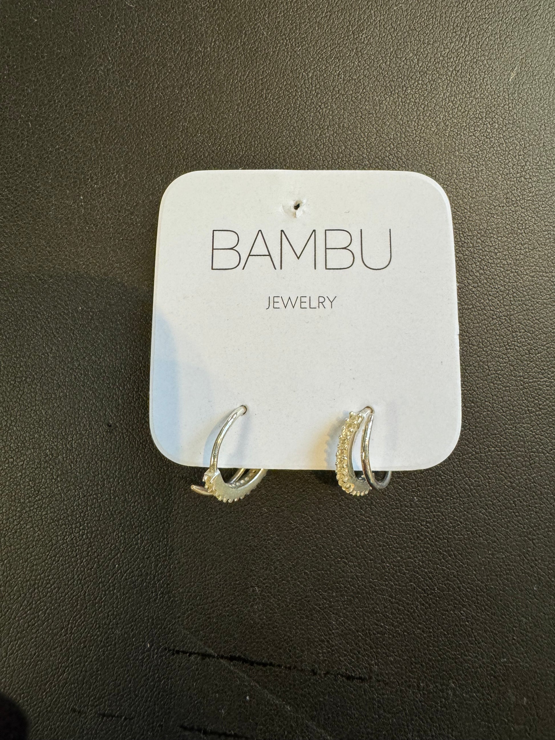 A pair of sleek Silver Embezzled Hoop Earrings from BAMBU Jewellery, adorned with a row of small, sparkling stones, are showcased on a square white card. The card prominently features the brand name "BAMBU" with "JEWELLERY" below it. These elegant earrings stand out against the textured black background due to their intricate detailing.