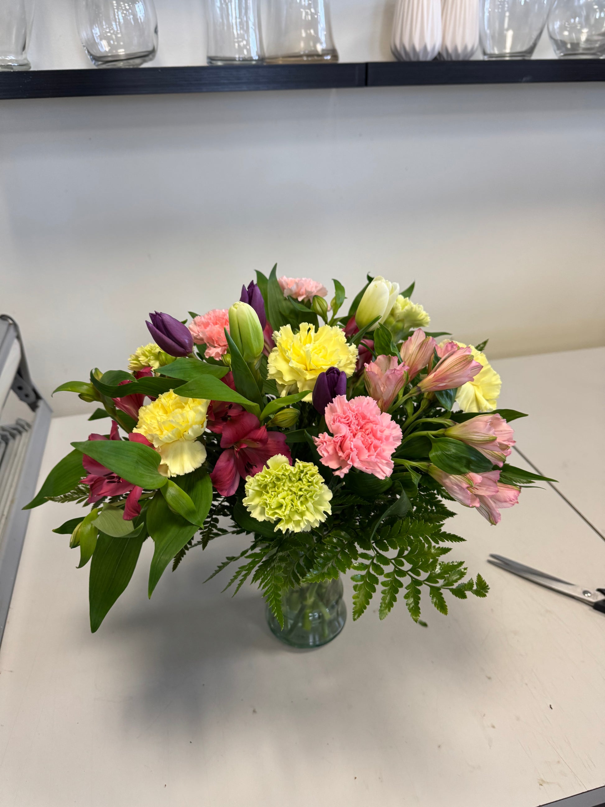 The Colours of Traditions Vase by Posies Flowers & Fashion showcases a vibrant mix of pink, yellow, green, and purple blooms with assorted leaves in a glass vase. Due to availability, some flower substitutions may occur.