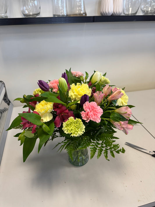 The Colours of Traditions Vase by Posies Flowers & Fashion showcases a vibrant mix of pink, yellow, green, and purple blooms with assorted leaves in a glass vase. Due to availability, some flower substitutions may occur.