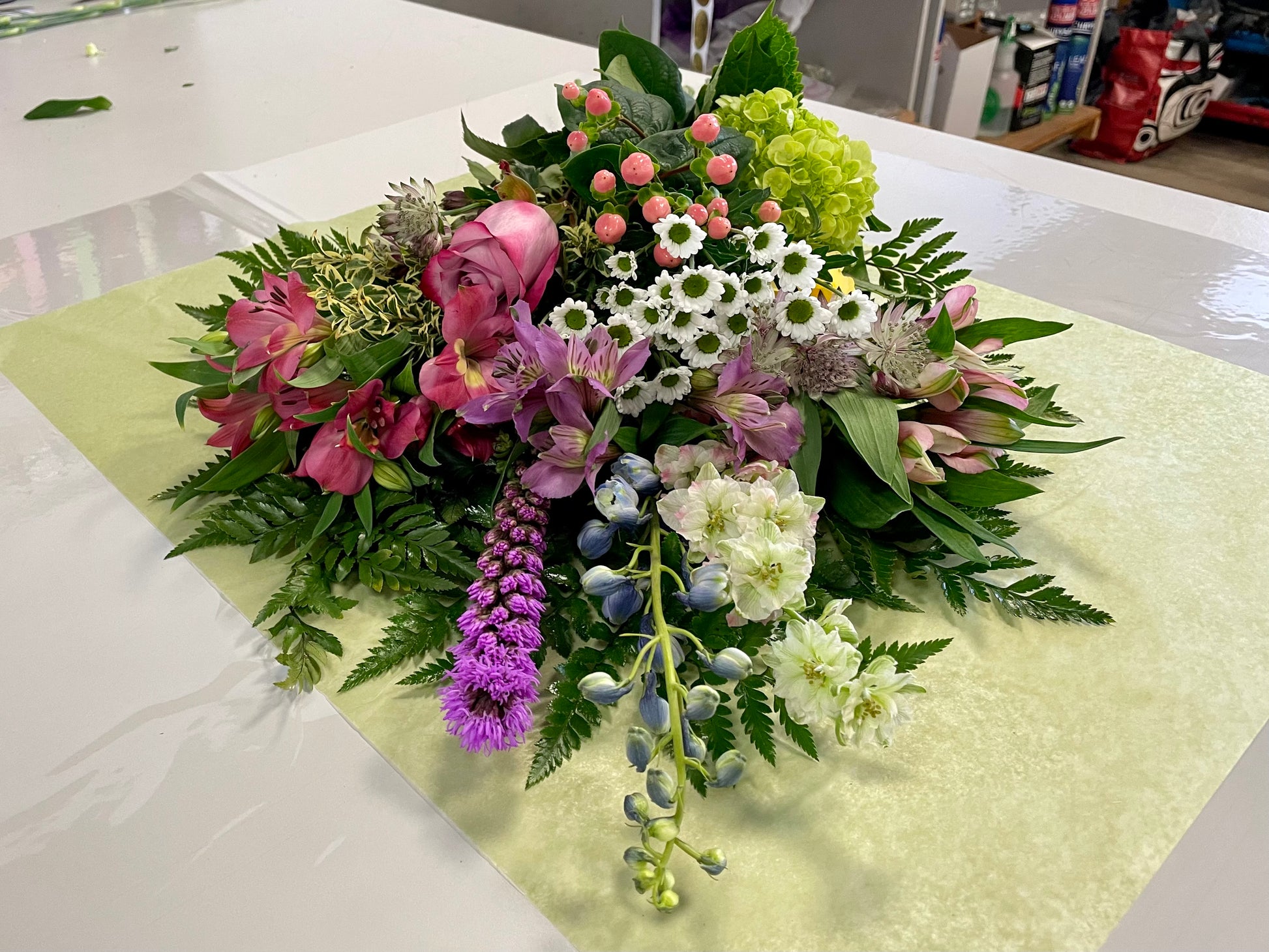 A stunning bouquet called Designer’s Choice Freshcuts from Posies Flowers & Fashion is resting on a white surface. The arrangement features vibrant orange and pink roses, cheerful yellow chrysanthemums, elegant white flowers, and assorted green foliage. It is partially wrapped in brown paper and elegantly tied with a burgundy ribbon.