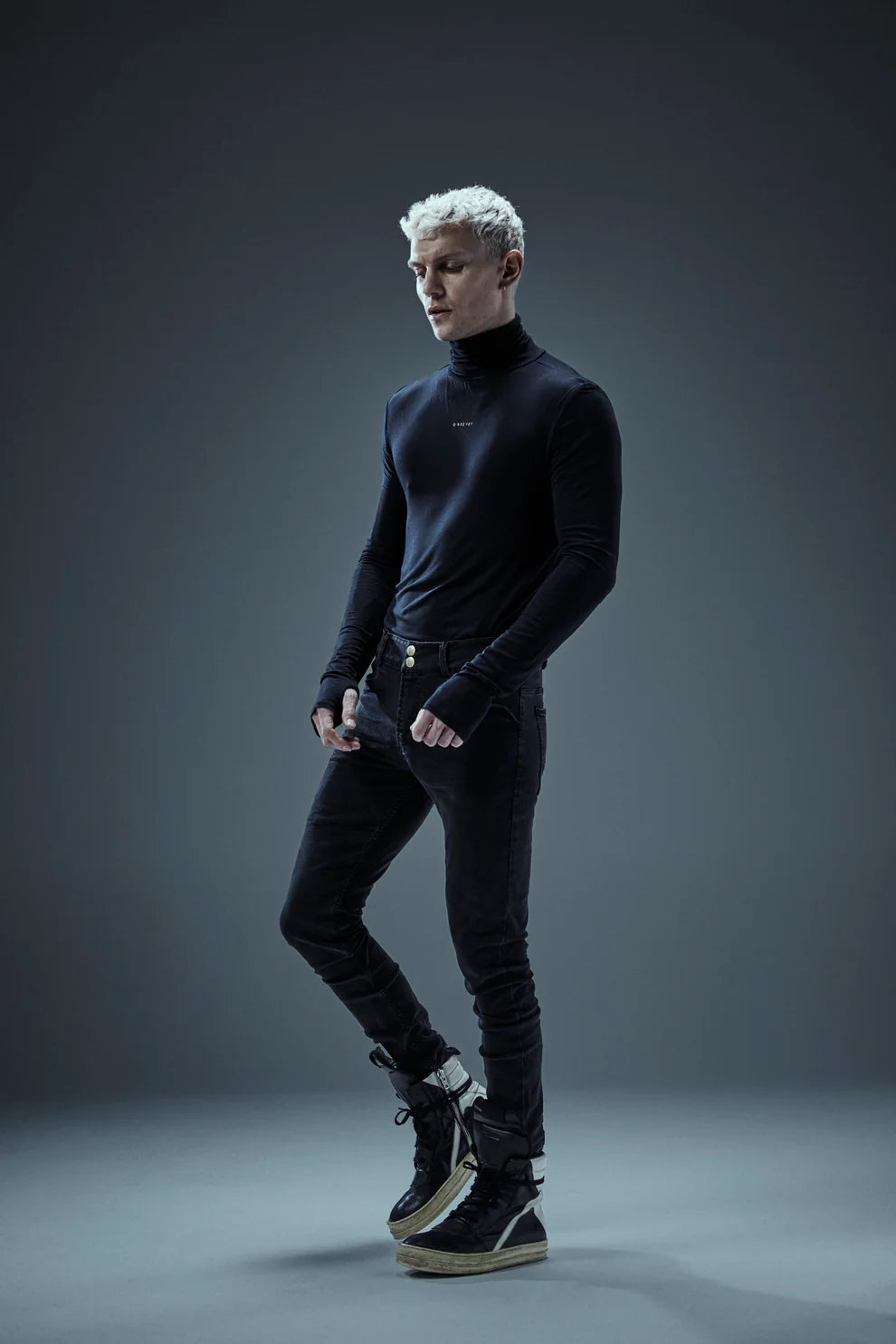 A person with short, light-colored hair stands confidently against a dark gray background. They are wearing a black turtleneck, Shuuto 2 Black DNM Skinny Jeans by Once We Were Warriors, and black high-top sneakers. Their fists are clenched at their sides as they look straight ahead.