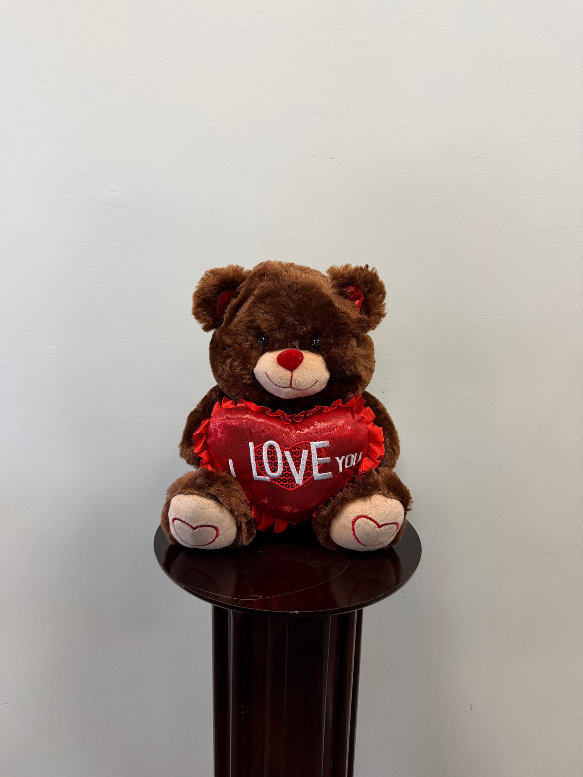 The “I Love You” Teddy Bear by Sproule sits atop a dark wooden pedestal, holding a red heart-shaped pillow with the words “I LOVE YOU,” against a plain light-colored wall.