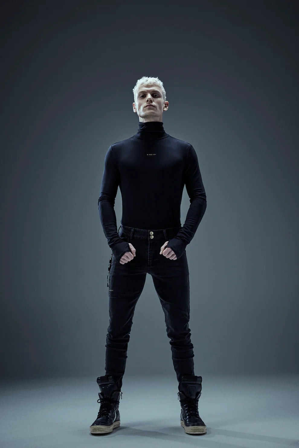 A person with short, light-colored hair stands confidently against a dark gray background. They are wearing a black turtleneck, Shuuto 2 Black DNM Skinny Jeans by Once We Were Warriors, and black high-top sneakers. Their fists are clenched at their sides as they look straight ahead.