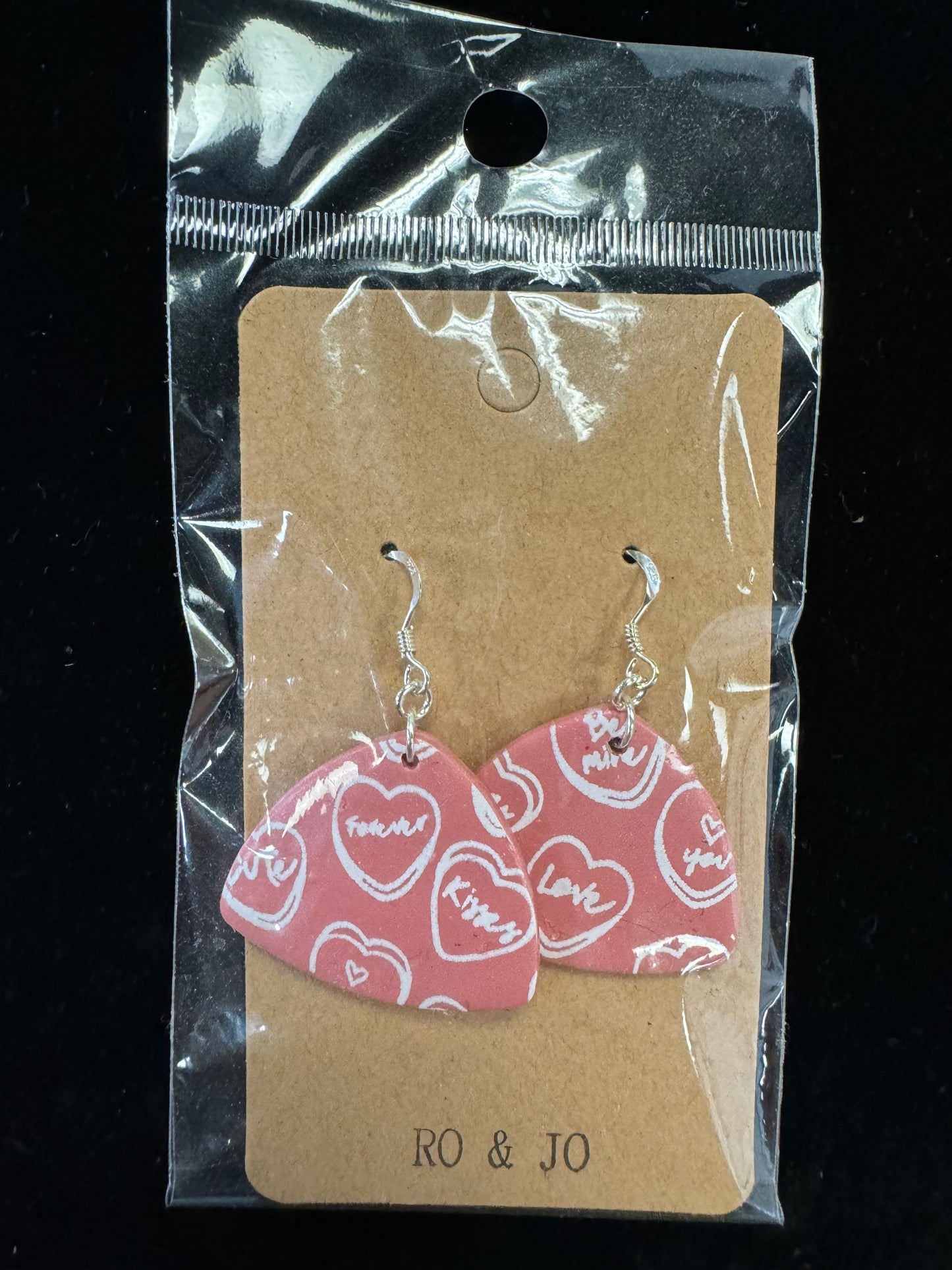 A pair of Ro&Jo Earrings 436, featuring a pink and white heart pattern, are displayed on a brown cardboard backing inside clear plastic packaging. These handmade earrings, designed with hooks for attaching to pierced ears, are beautifully showcased in packaging labeled "RO&JO" at the bottom, highlighting the charm of artisan jewelry.