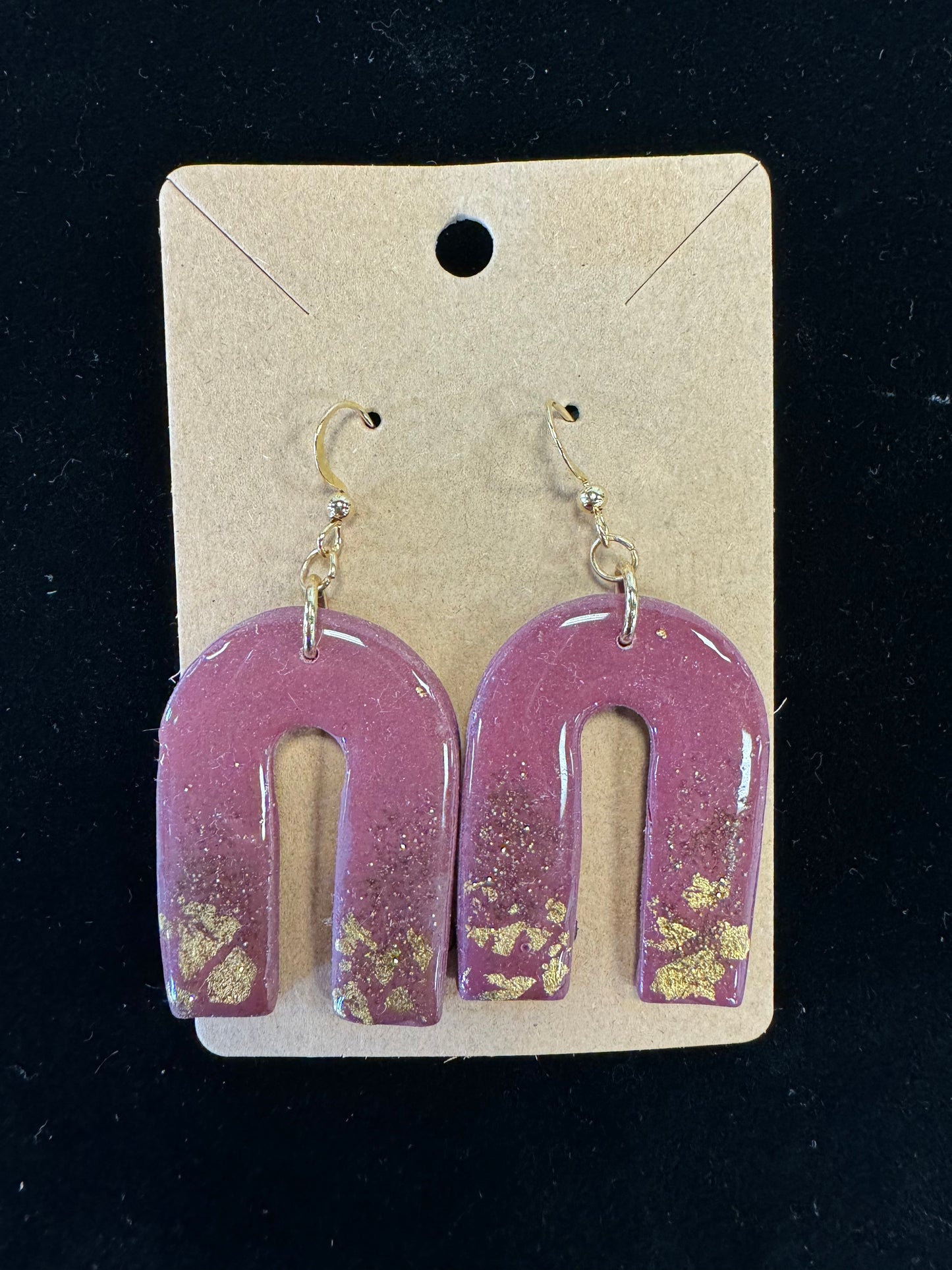 Ro&Jo Earrings 435 by RO&JO are handmade polymer clay earrings, U-shaped with a glossy pink finish and gold fleck accents. They are attached to a simple brown card with a hole at the top for hanging and feature gold-colored hooks for wearing. The background is black.