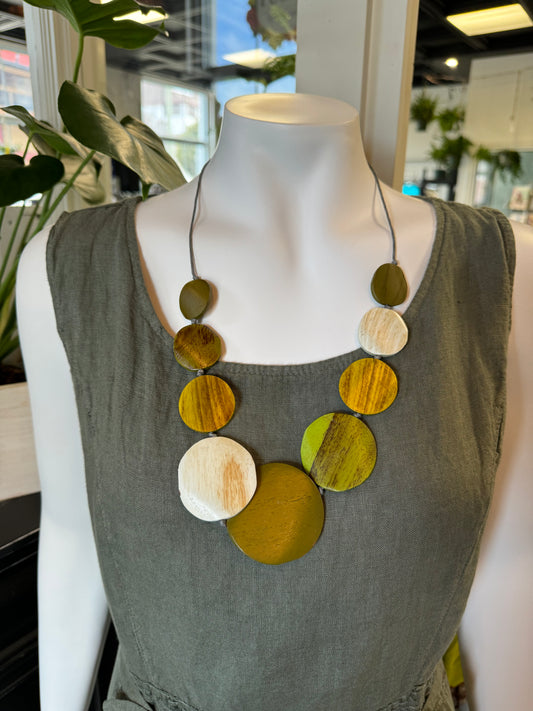 A mannequin dressed in a sleeveless, green, textured dress is adorned with the Green Circle Necklace by Suzie Blue, featuring multi-sized charms in shades of green, brown, and white, arranged asymmetrically. The background evoking an Indonesian ambiance contains lush plants and shop interior elements.