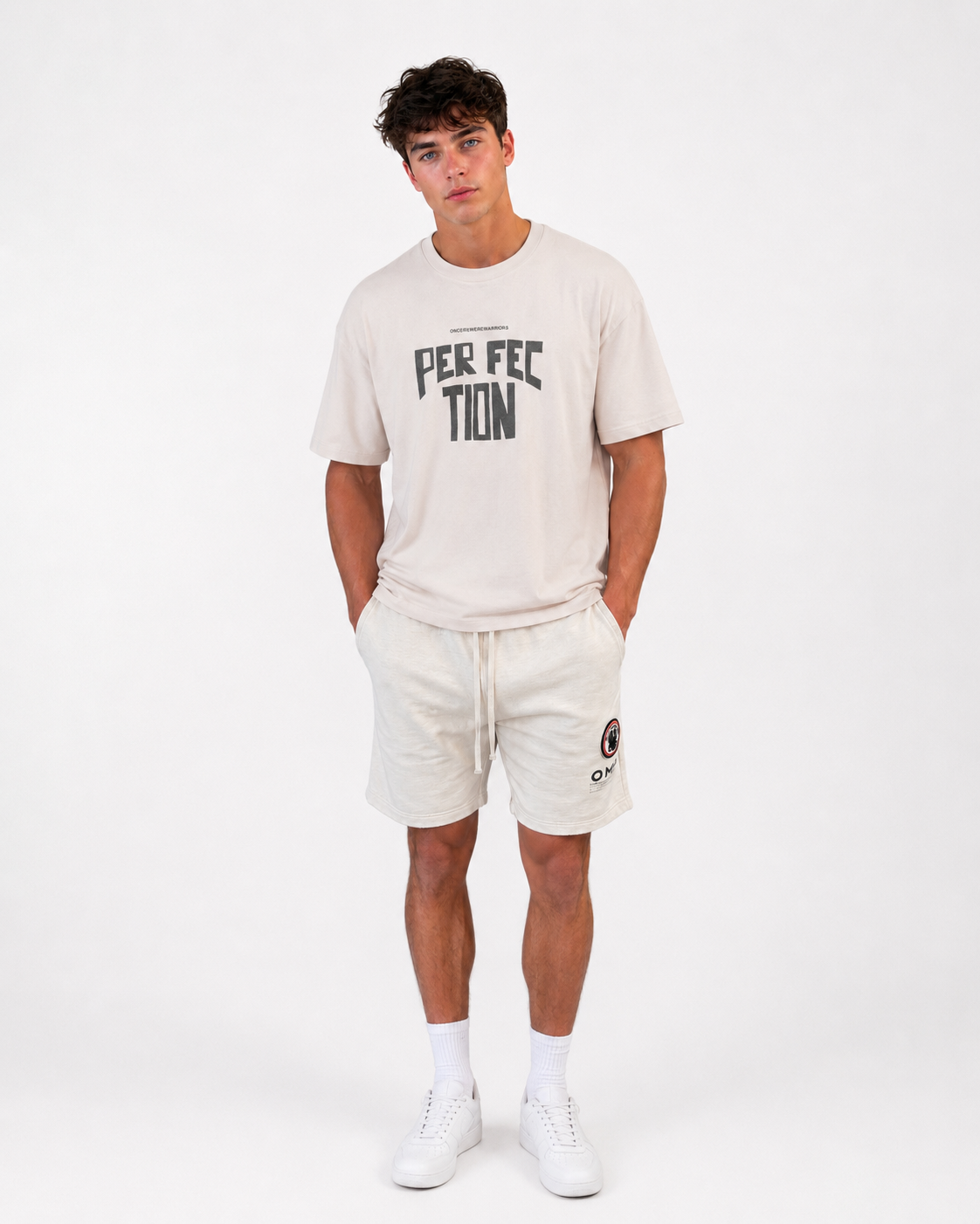 A man stands against a white background wearing the Once We Were Warriors Talan Tee - WHITE SAND with "PERFECTION" text, paired with light shorts, white socks, and sneakers. His hands are in his pockets and his expression is neutral.
