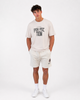 A man stands against a white background wearing the Once We Were Warriors Talan Tee - WHITE SAND with "PERFECTION" text, paired with light shorts, white socks, and sneakers. His hands are in his pockets and his expression is neutral.