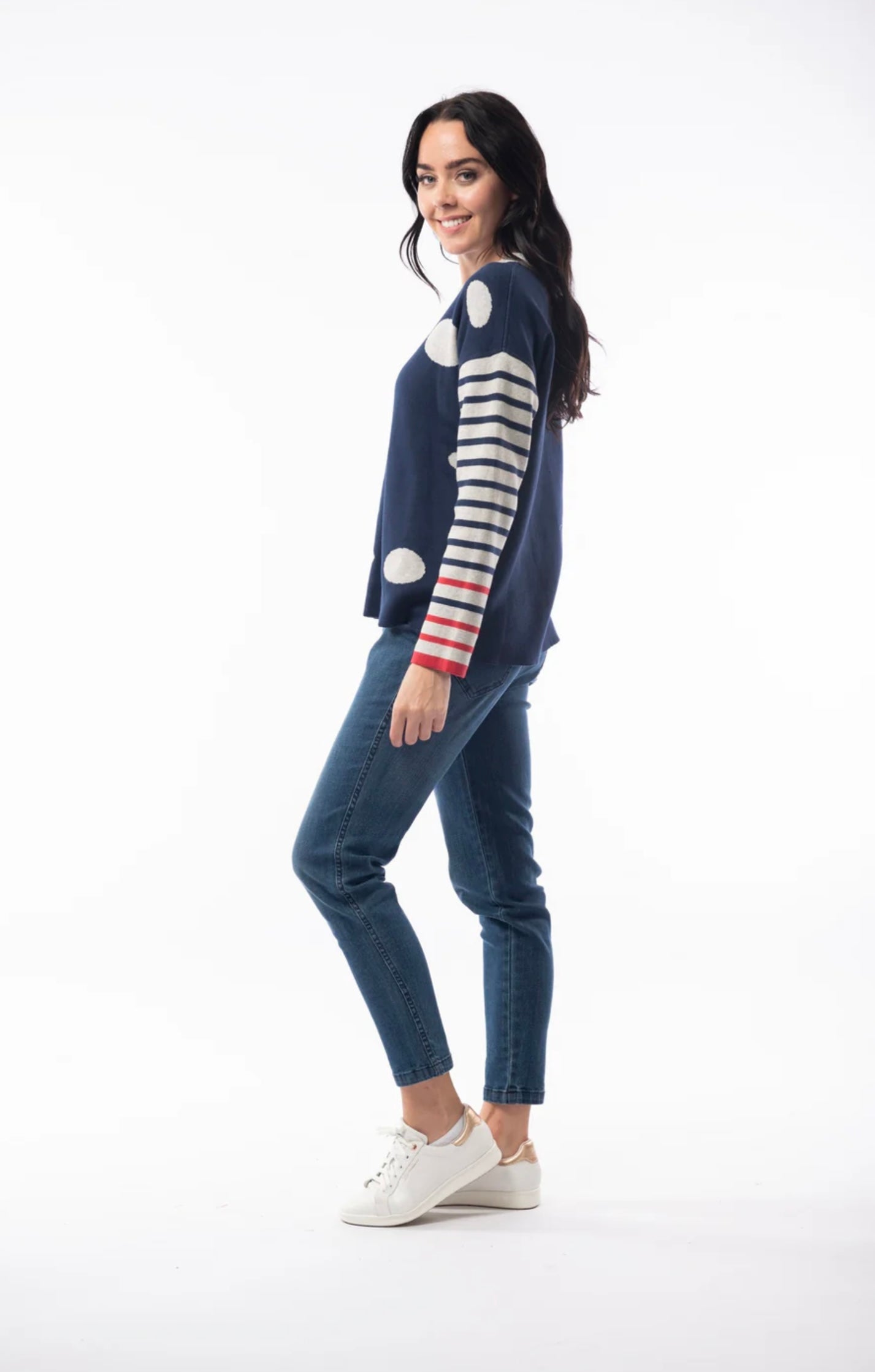 A woman with long dark hair is wearing the ORIENTIQUE - REVERSIBLE ROUND NECK SPOT/STRIPE JUMPER (1229) made of organic cotton, featuring a navy blue background adorned with white polka dots and red striped sleeves. She's paired it with blue jeans and white sneakers, and stands in a casual pose, slightly leaning back with her hands behind her. The background is plain white.