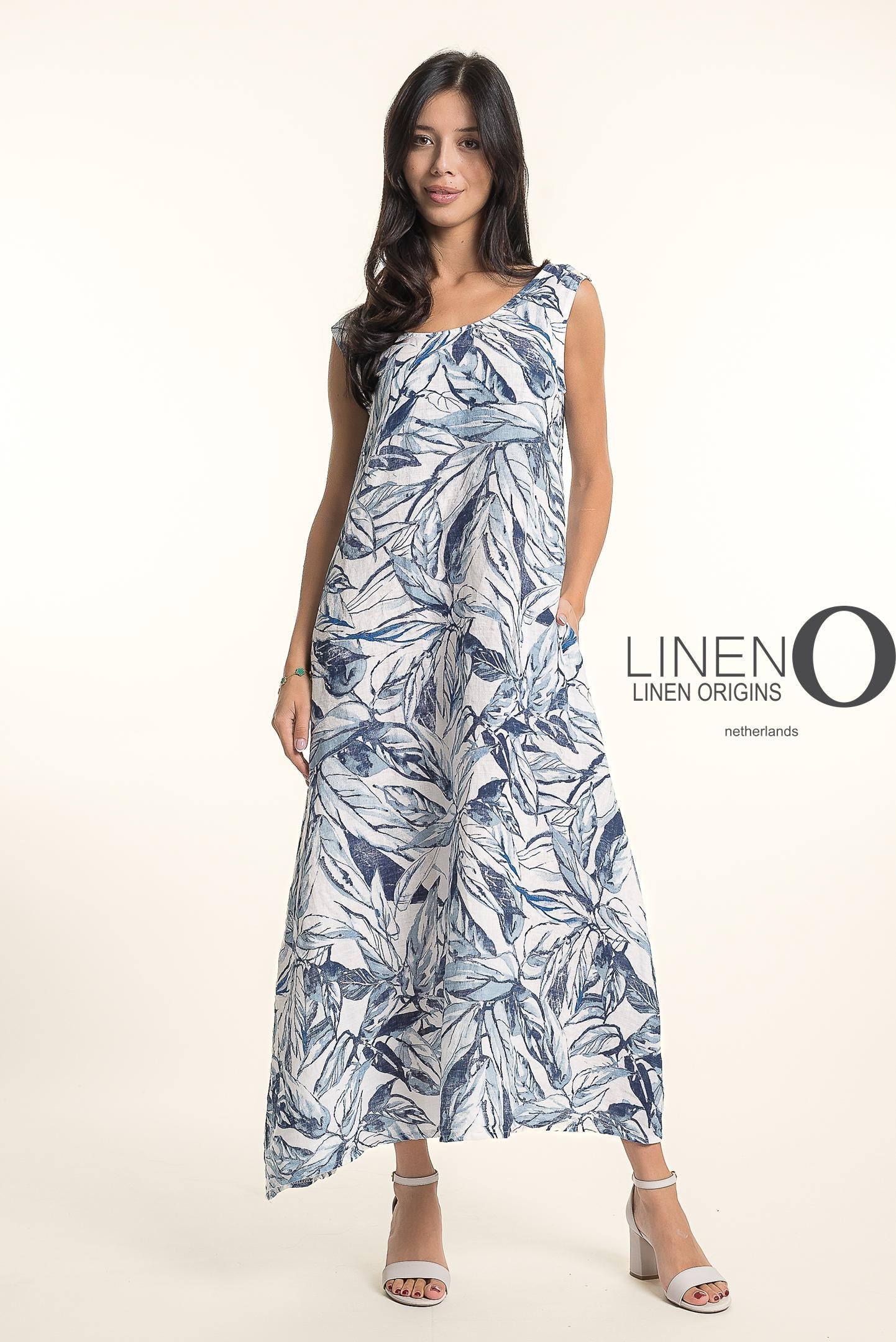 A woman with long dark hair wears white heeled sandals and the sleeveless, ankle-length French Linen Dress TP1720P-11 Bianca, featuring a blue and white leaf pattern by LinenO. "LINEN O" appears beside her against a light background.