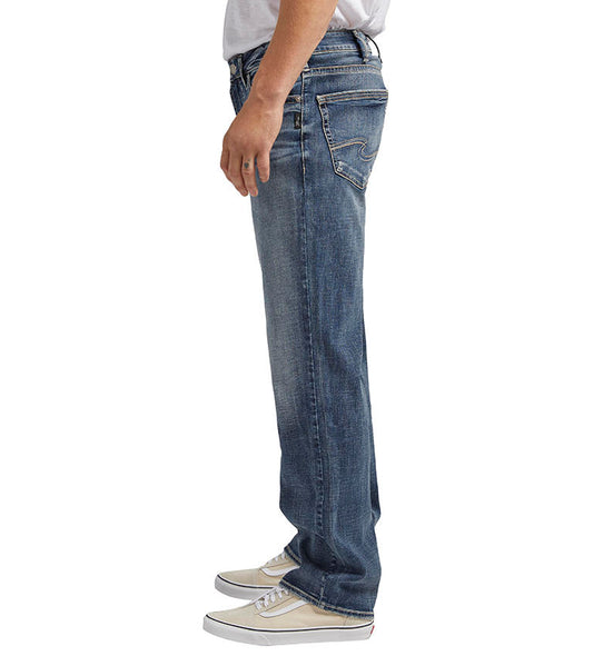 A person wearing a white t-shirt, blue jeans, and white sneakers is standing and facing sideways to the right. Only the lower two-thirds of the body are visible in the image. The Grayson Classic Fit Straight Leg Jeans from Silver Jeans Co. feature a relaxed fit with slightly faded detailing in a dark indigo wash.