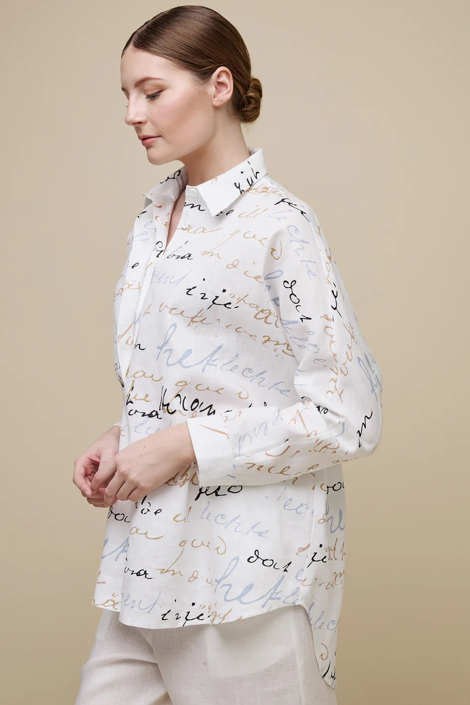 A person stands against a beige background, wearing the UCHUU Tunic Style Blouse (CS24-632) with handwritten-style text patterns. They pair it with loose white pants and black sandals. Their right hand rests in the pant pocket, and they have a neutral expression.