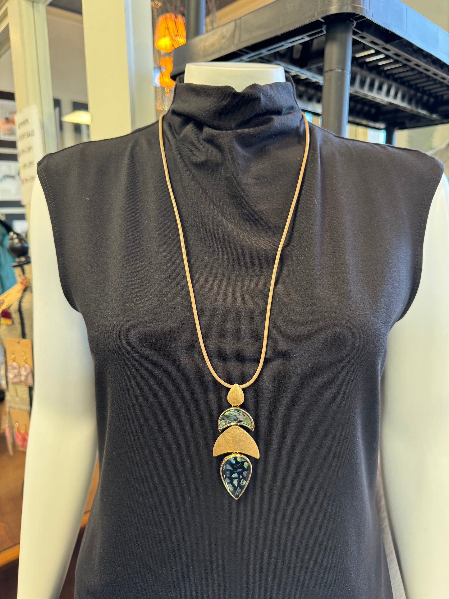 A mannequin displays a black sleeveless top paired with a striking fashion accessory—a long gold necklace from Elise Accessories, named Necklace 2, featuring three teardrop-shaped pendants with green and black accents. Set in a clothing store, it seamlessly blends into the jewelry collection on display amidst an array of garments.