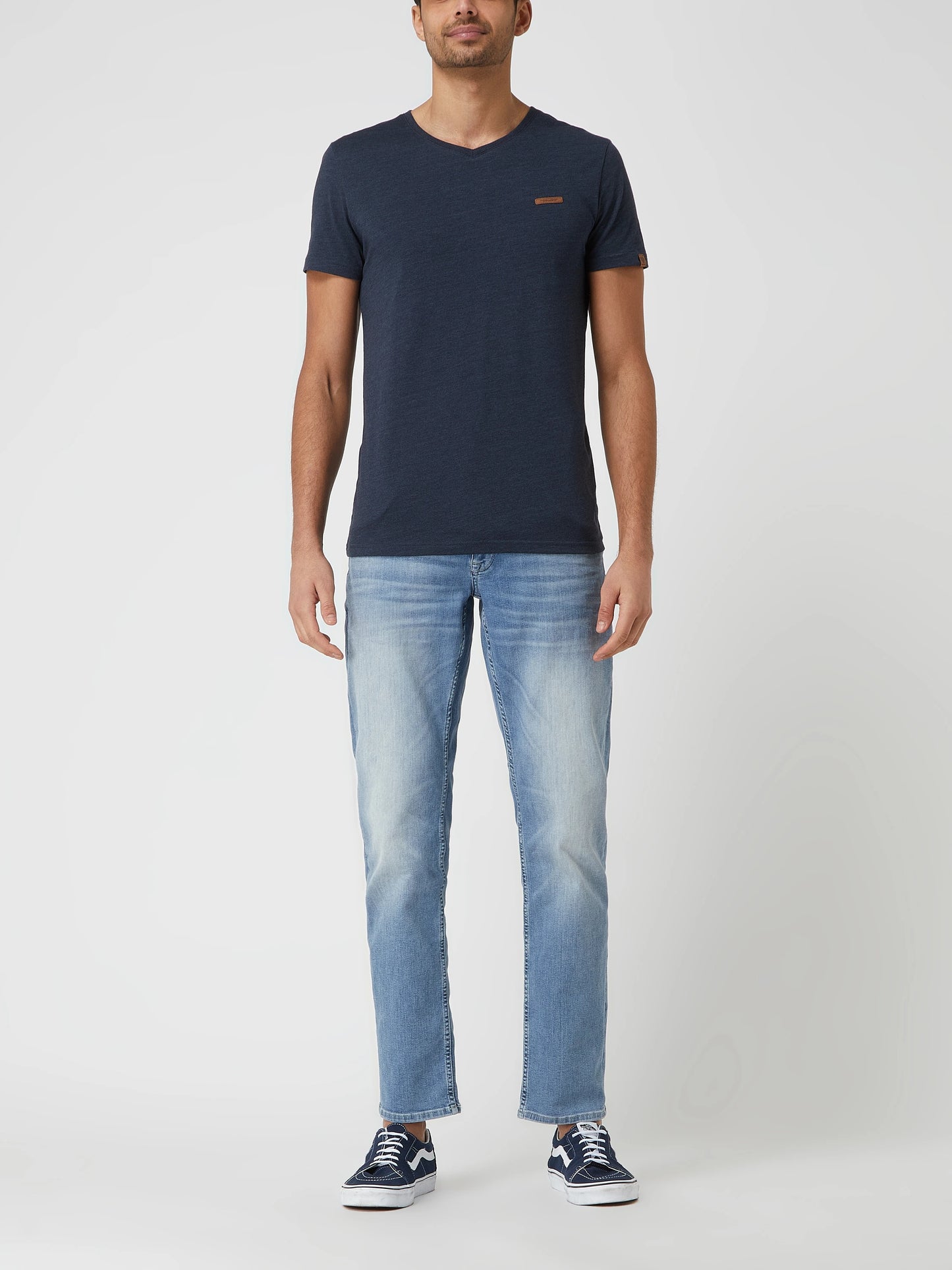 A man with short brown hair is wearing a beige pair of pants and a plain navy blue V-neck t-shirt from RAGWEAR's VENIE collection. He stands facing the camera against a plain white background, with a neutral expression and his arms relaxed by his sides.