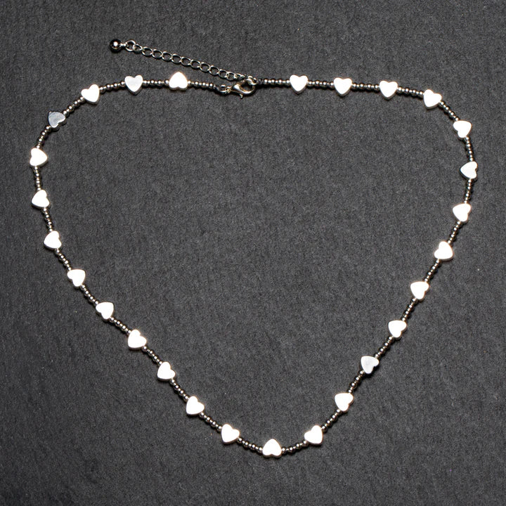 A delicate and ethically sourced Short Heart Necklace In Silver Plate by Suzie Blue features small, shiny heart-shaped charms evenly spaced along a thin chain. The charms are arranged in a slight curve against a dark, textured background, and the chain is finished with an adjustable clasp and a small dangling bead.