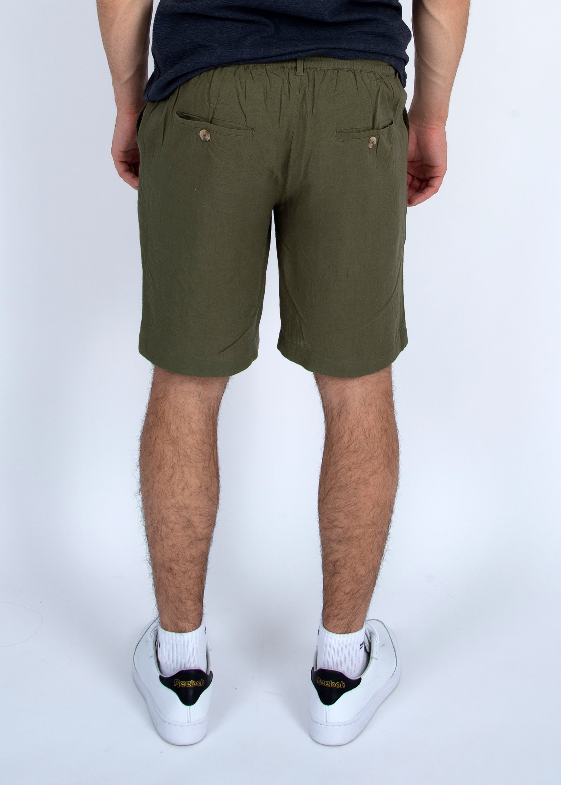 A person faces away, wearing HEDGE Linen Blend Shorts Vine 41MW507S in olive green with two buttoned back pockets, a dark t-shirt, and white sneakers with black and yellow accents against a plain light background.