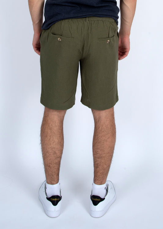 A person faces away, wearing HEDGE Linen Blend Shorts Vine 41MW507S in olive green with two buttoned back pockets, a dark t-shirt, and white sneakers with black and yellow accents against a plain light background.
