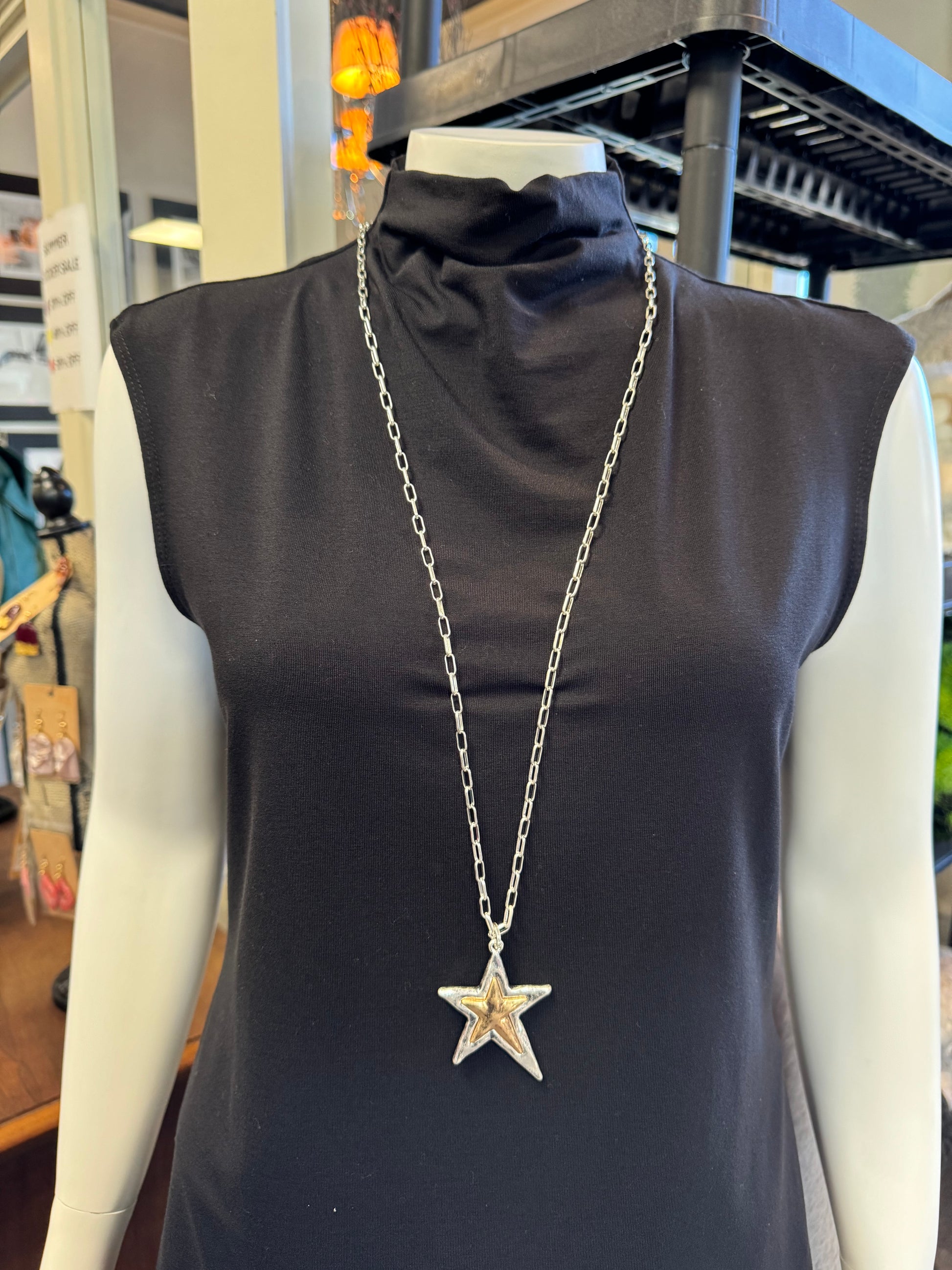 A mannequin showcases a black sleeveless turtleneck shirt, enhanced by a striking piece from Elise Accessories: Necklace 6. This long silver necklace, featuring a prominent star pendant, captivates against the backdrop of blurred shelving and miscellaneous items.