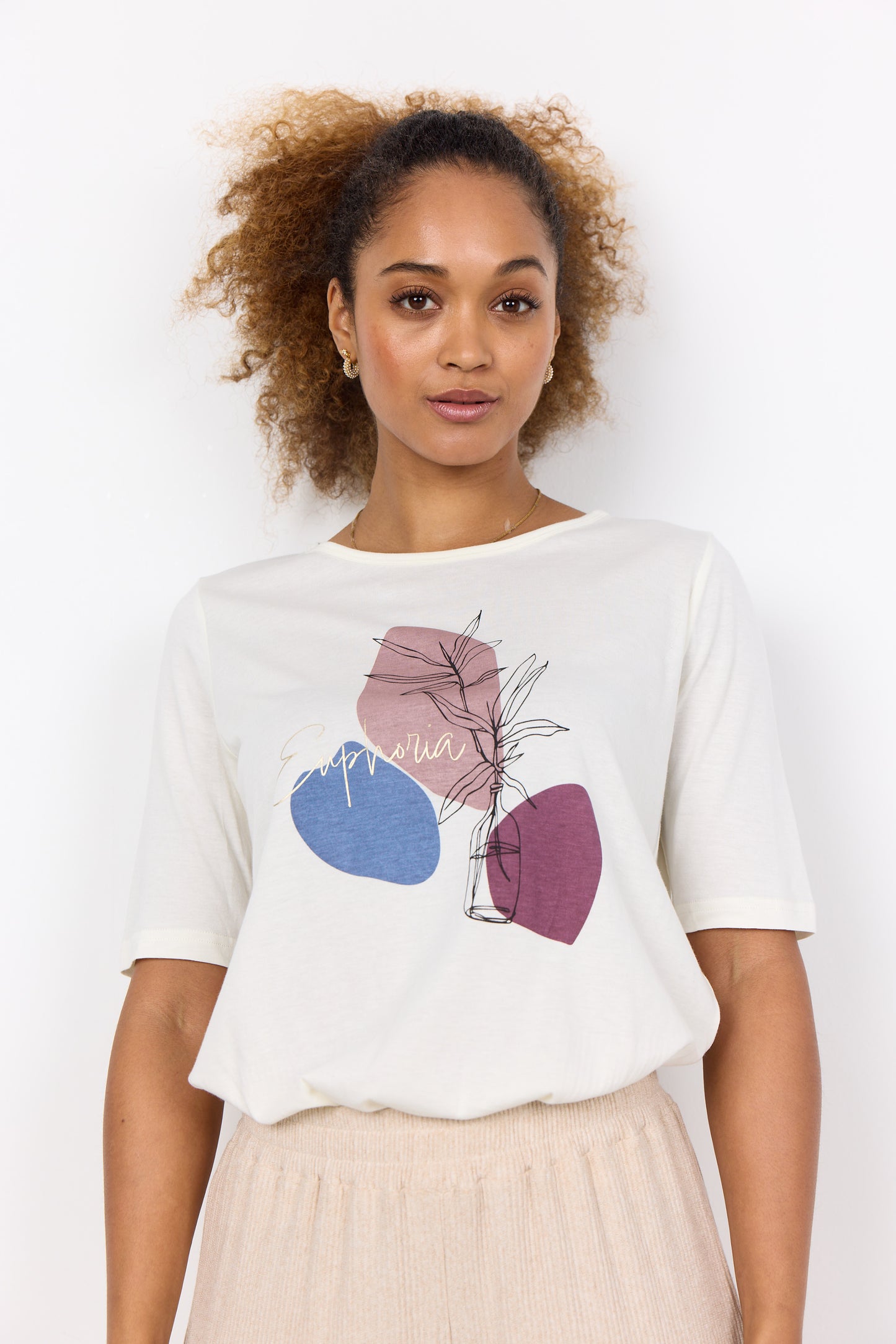 A woman with curly hair pulled back, wearing the stylish T-Shirt Felicity Woodrose SC-FELECITY FP 475 from RAGWEAR featuring abstract shapes and the word "Euphoria," stands in front of a white background. She pairs it with light-colored pants, making her look effortlessly chic.