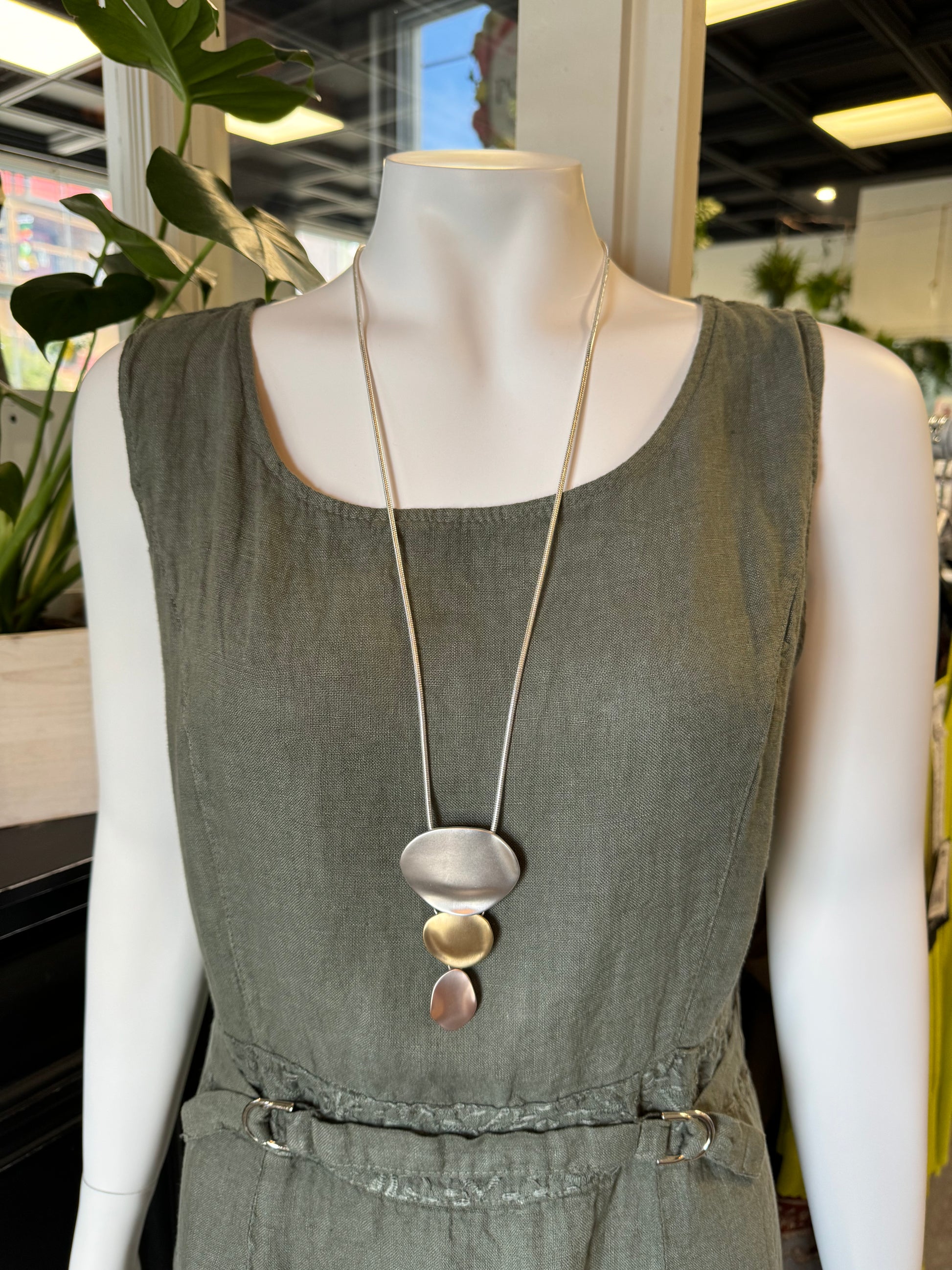 A white mannequin dressed in a sleeveless, green, linen dress cinched at the waist is displayed. The trendy look is accessorized with the Posies Flowers & Fashion Inc. Rose Gold, Silver and Gold Necklace featuring three abstract pendant shapes. The background includes indoor plants and shop decor.