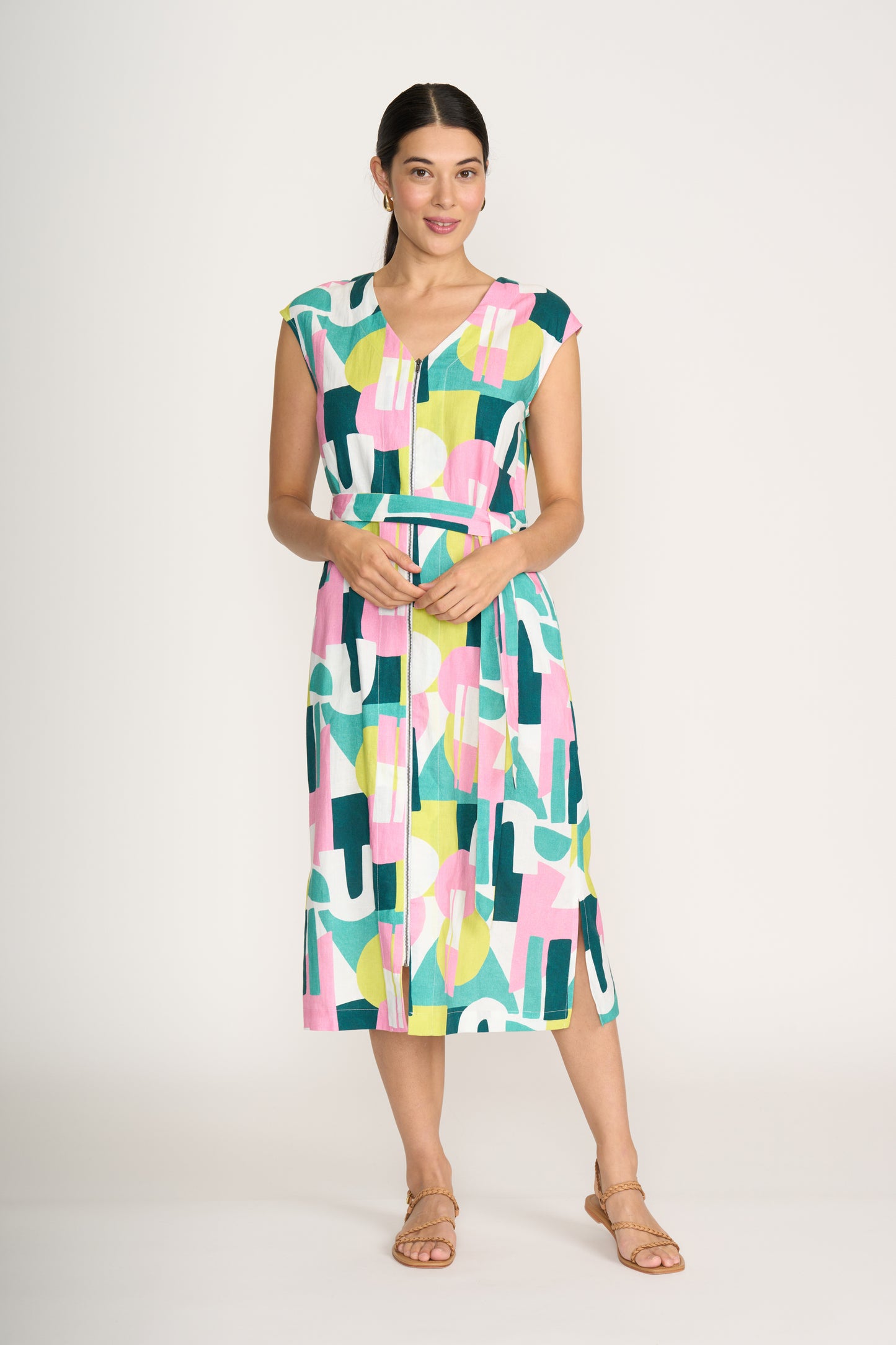 A woman wears the FOIL You Little Zipper Dress Spectrum FO8294—a sleeveless, belted midi dress with a geometric pink, green, yellow, and white pattern—paired with tan sandals, standing against a plain white background.