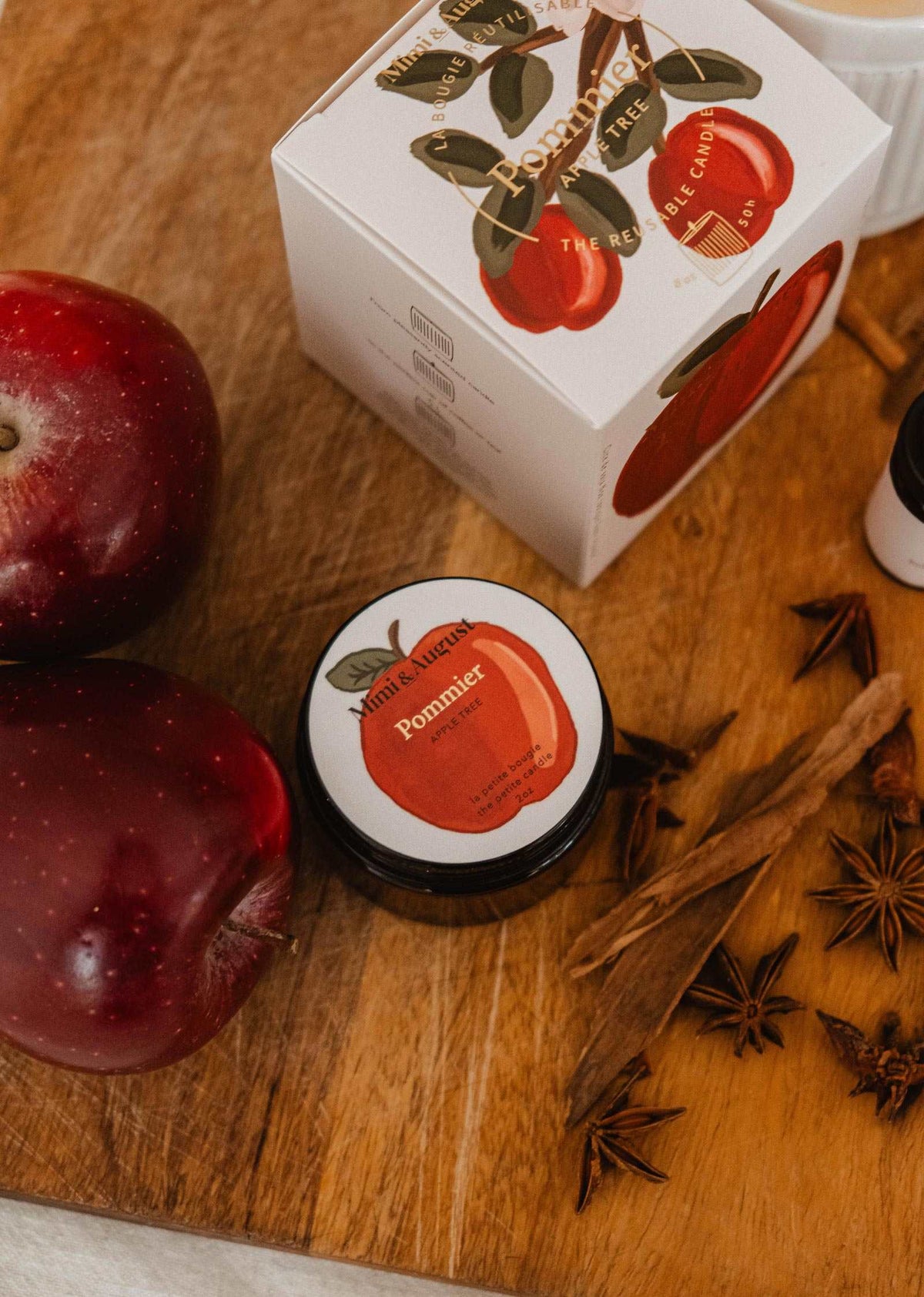 A 2 oz jar of Mimi & August’s Pommier soy candle, featuring an apple illustration, sits on a wooden surface alongside two red apples, a branded box, star anise, cinnamon sticks, and a small bottle—creating an inviting display for this delightful apple cinnamon scent.