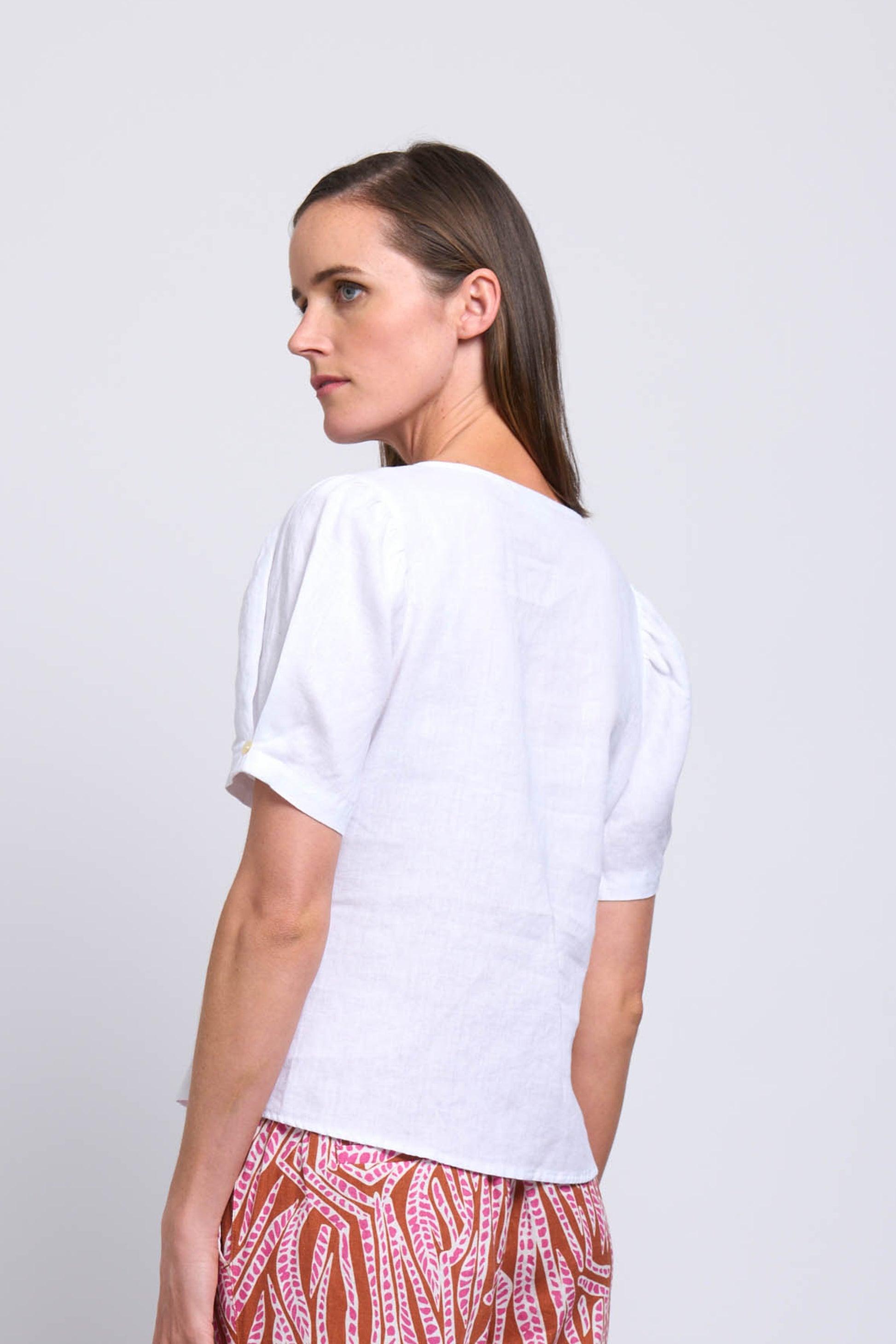 A woman with straight brown hair models the FOIL Good Tuck Shirt FO7800 in white, standing sideways and looking over her shoulder against a plain light background.