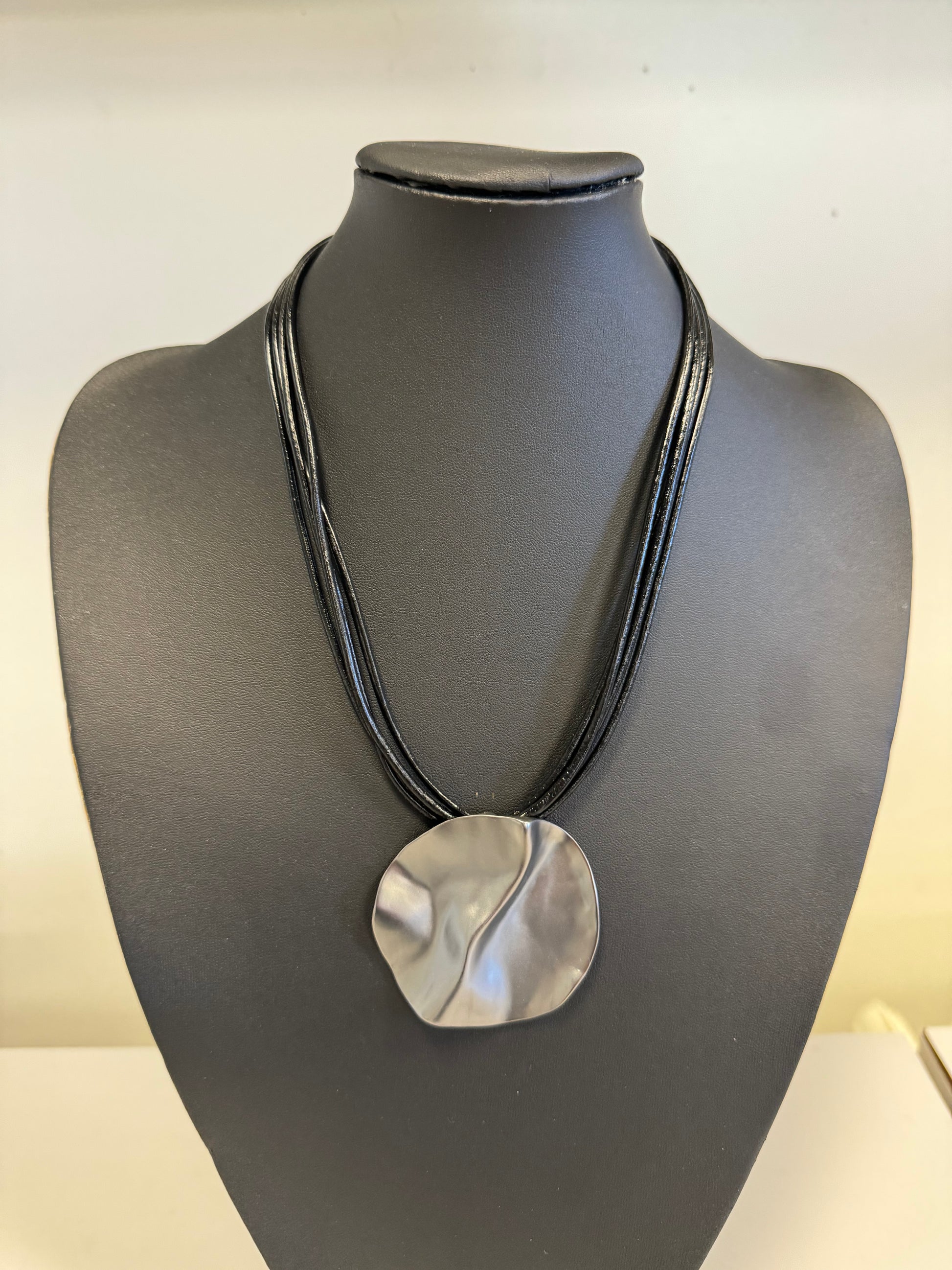 The Roxanne 29 Necklace by Elise Accessories is a striking fashion piece, featuring a pendant with a wavy metallic finish. It elegantly hangs from multiple thin black cords, creating a layered look. This necklace, perfect for any jewelry collection, is displayed against a plain light-colored background.