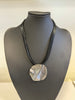 The Roxanne 29 Necklace by Elise Accessories is a striking fashion piece, featuring a pendant with a wavy metallic finish. It elegantly hangs from multiple thin black cords, creating a layered look. This necklace, perfect for any jewelry collection, is displayed against a plain light-colored background.