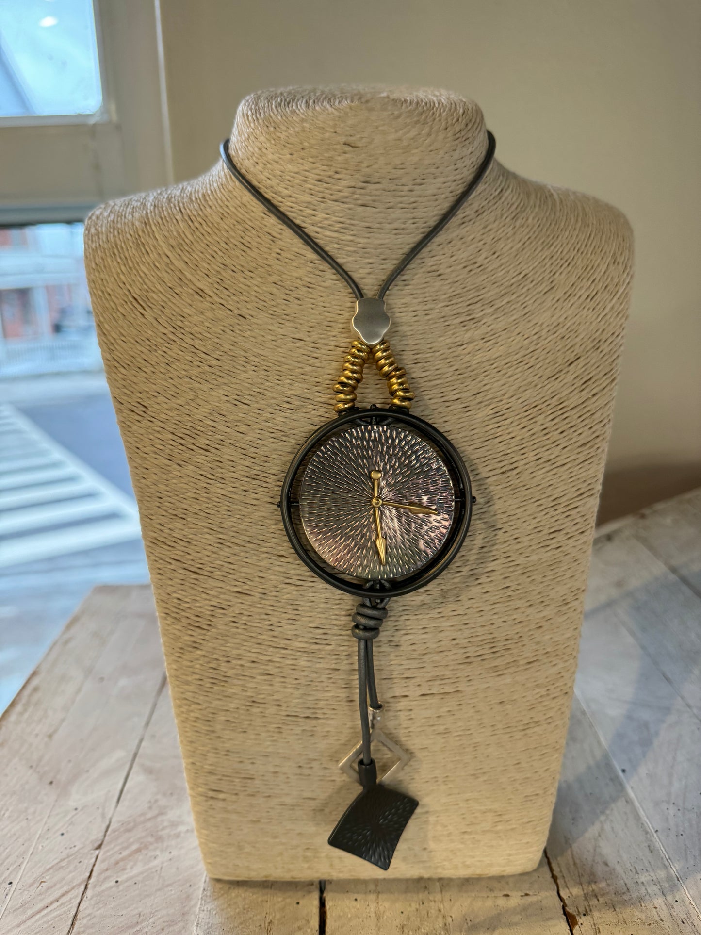 The Clock Necklace by Posies Flowers & Fashion Inc. is elegantly showcased on a beige, textured stand. This stunning piece features a large, round pendant with a textured metallic surface and gold-colored details. The pendant hangs from a black cord adorned with decorative gold beads and finishes with a black tassel at the bottom.