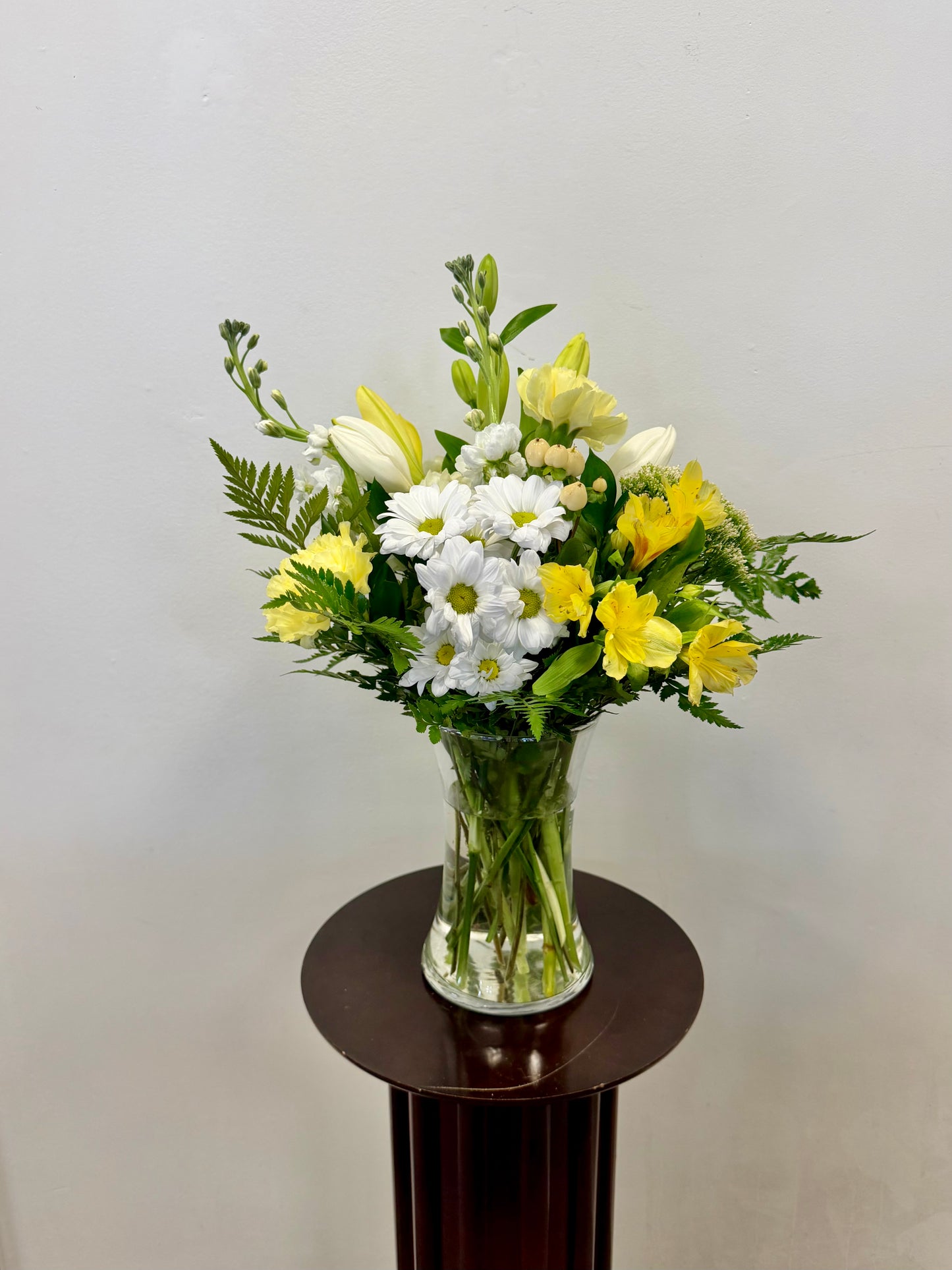 The Posies Flowers & Fashion Whites and Yellows Vase, featuring a fresh mix of white and green flowers with foliage in a clear glass vase, sits on a small round dark wooden table against a plain light-colored wall.