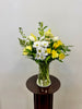 The Posies Flowers & Fashion Whites and Yellows Vase, featuring a fresh mix of white and green flowers with foliage in a clear glass vase, sits on a small round dark wooden table against a plain light-colored wall.