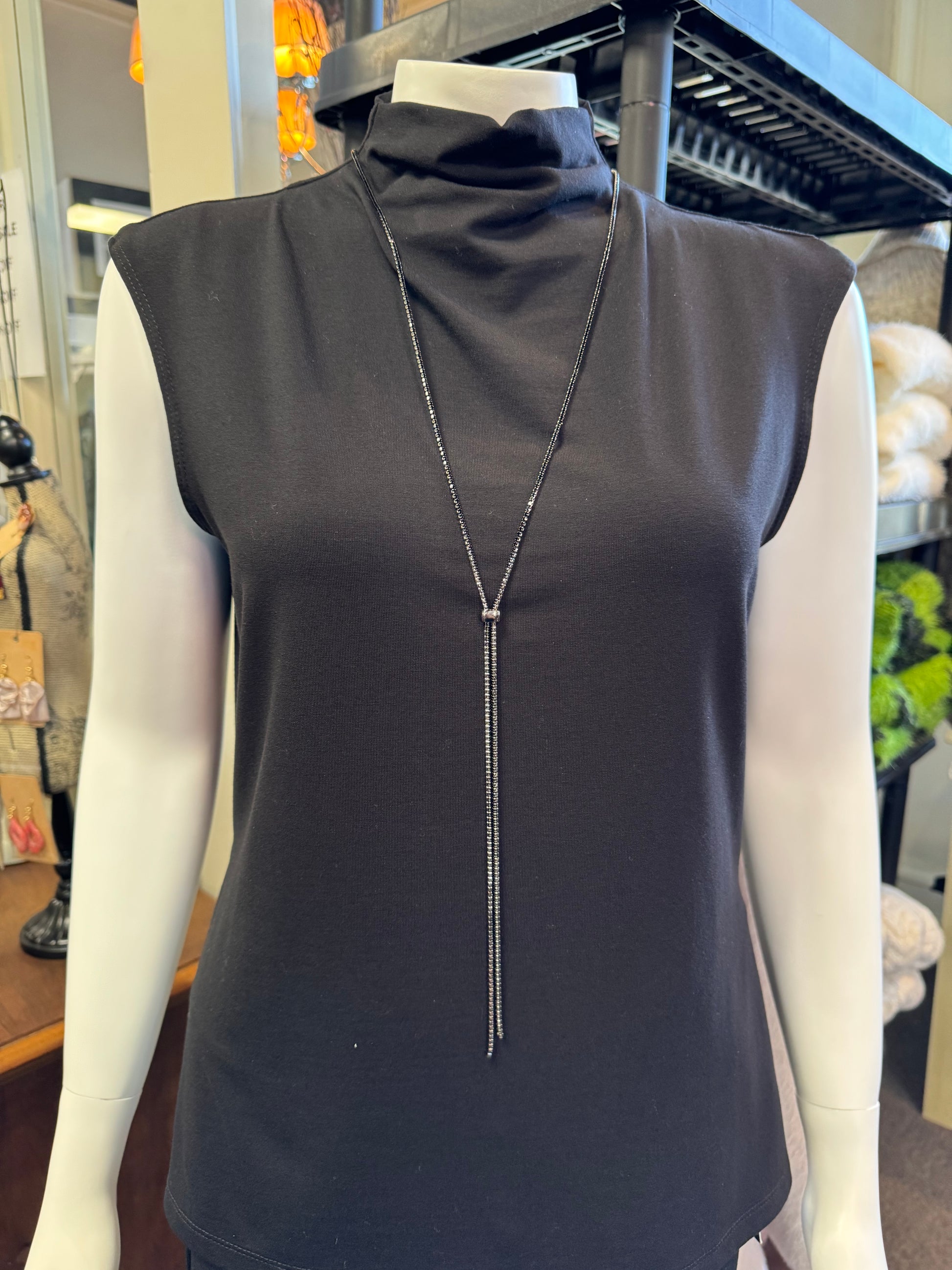 A mannequin is elegantly dressed in a sleeveless black turtleneck top, highlighted by the Posies Flowers & Fashion Inc. Sparkly Necklace. This long silver Y-shaped piece shimmers with stunning crystals, set against a backdrop of diverse fabrics and decorative elements in a store setting.