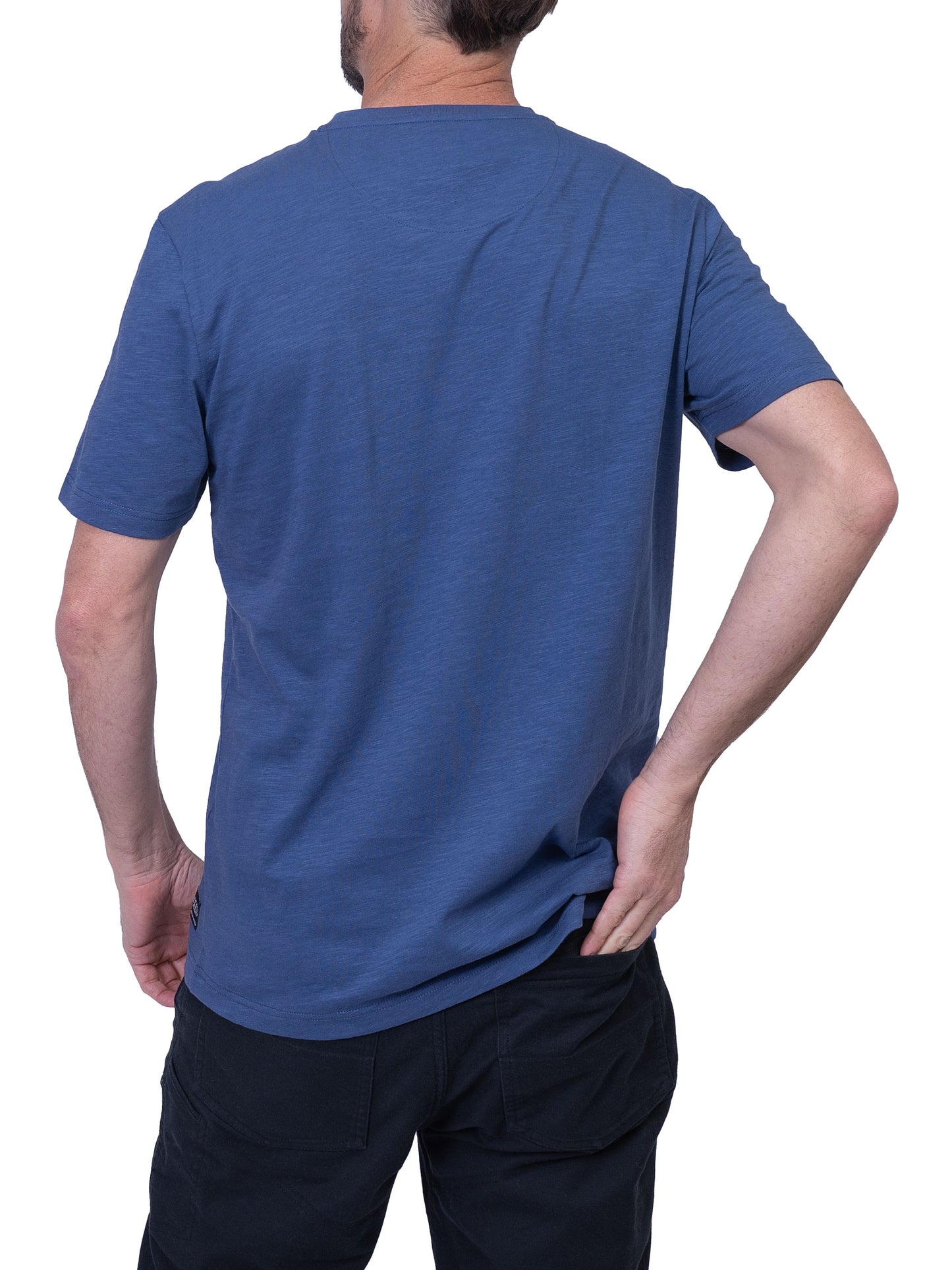 A person stands wearing the Men’s Cotton T-Shirt - BLUE from Silver Jeans Co. The blue, short-sleeved T-shirt features a light blue front pocket with a floral pattern. They gaze to the side with one hand in their black pants pocket, embodying the epitome of casual style against a white background.