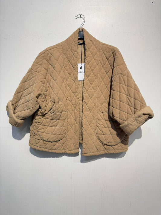 The NOELINE Quilted Jacket 2442-2 in Camel, featuring a tan quilted cotton design with wide sleeves, an oversized fit, and rounded pockets, hangs on a hanger against a plain white wall.