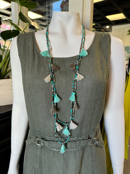 A mannequin wearing a sleeveless, olive green dress with a belted waist. Around its neck is the Beaded with Ruffles Necklace by Suzie Blue adorned with turquoise beads and tassels in teal, gray, and white. The background features indoor plants and a glimpse of a retail store’s interior.