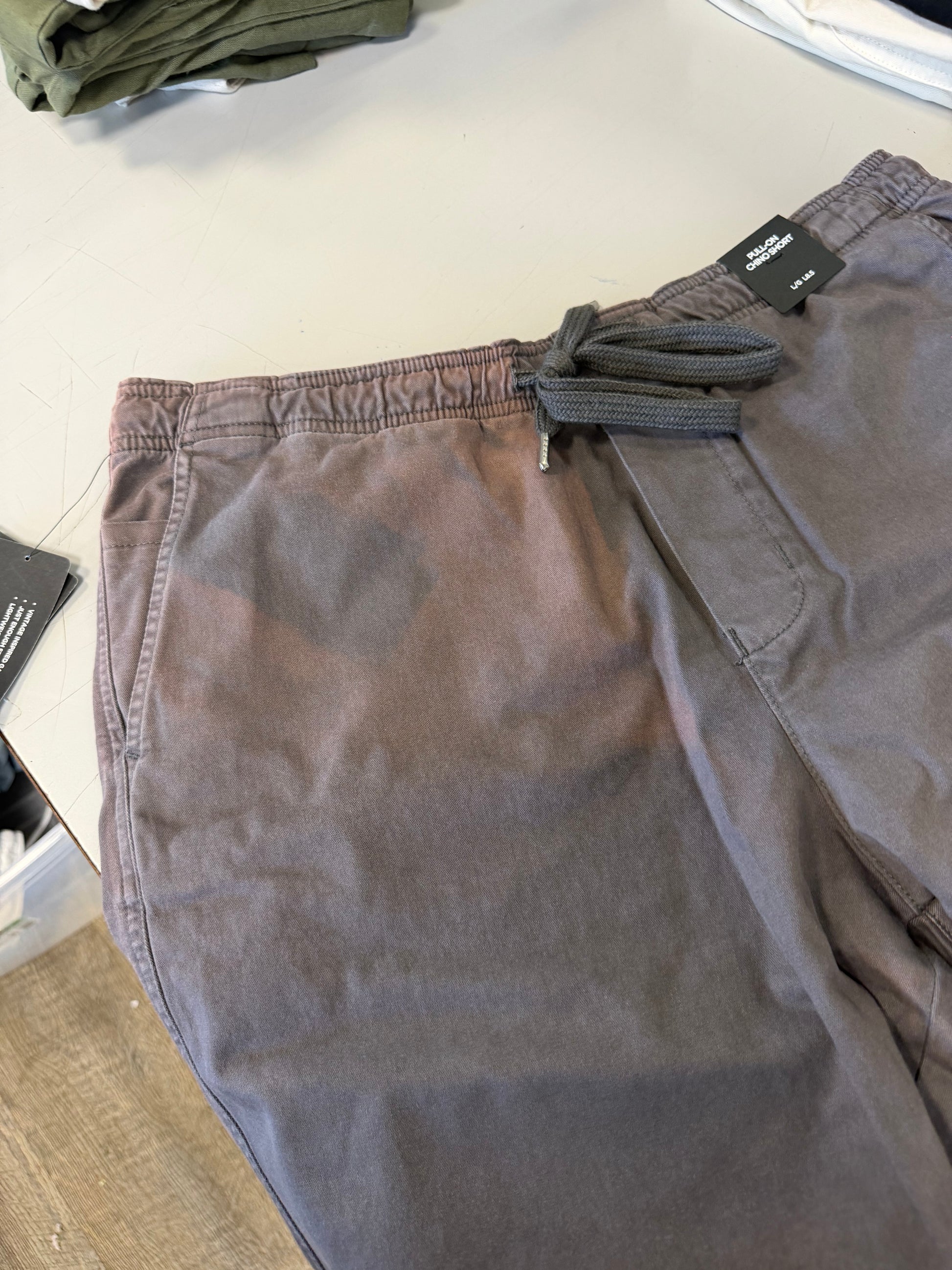 A pair of Silver Jeans Co. PULL-ON CHINO SHORT DARK GREY (Style M52155CBT639) with an elastic waistband and visible front pocket is displayed on a light surface, with folded clothes partially seen in the background.