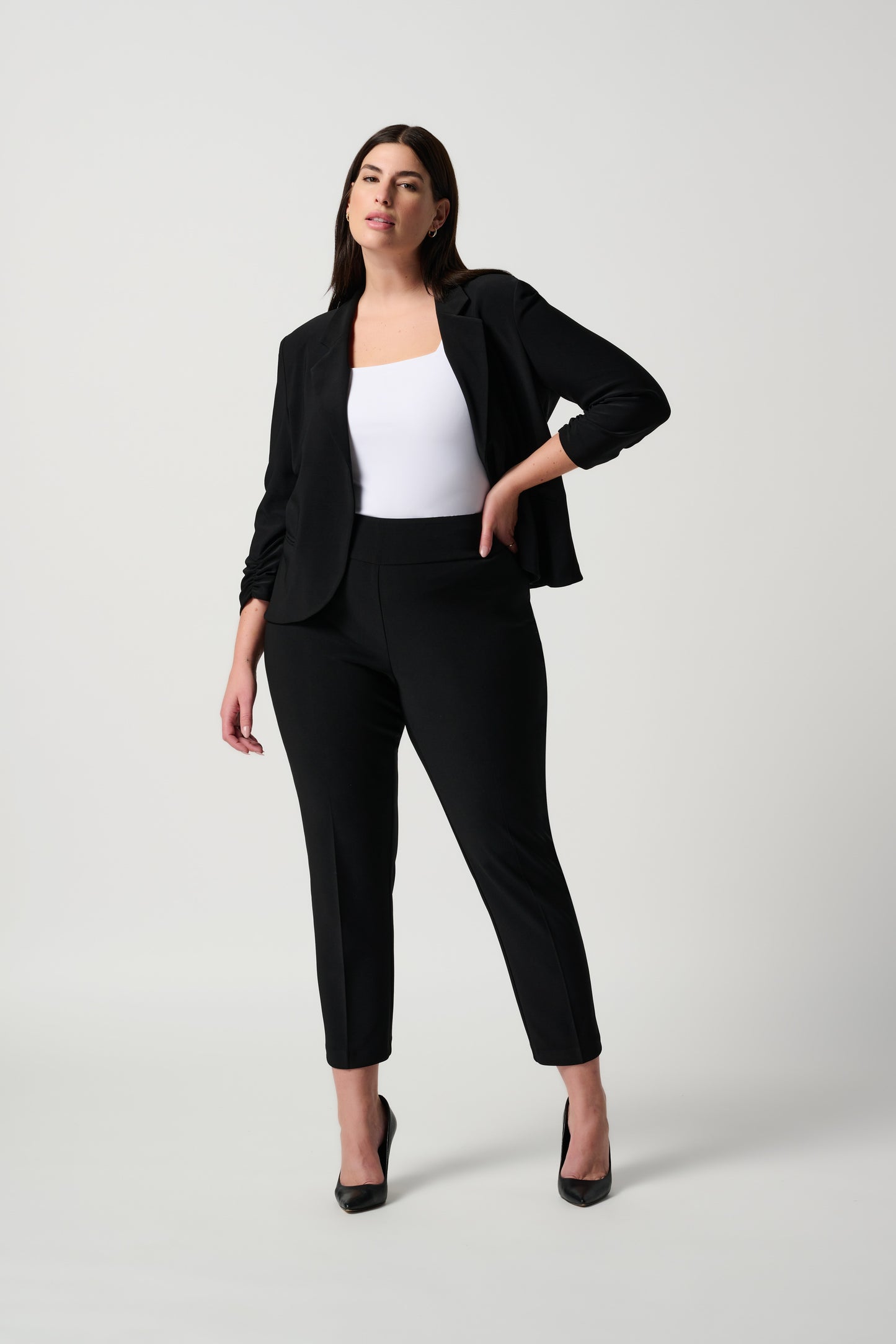 A person is donning Joseph Ribkoff's Classic Cropped Pant paired with black high-heeled shoes. The top portion of their outfit is a white shirt, and their arms rest by their sides. The background is plain and white.