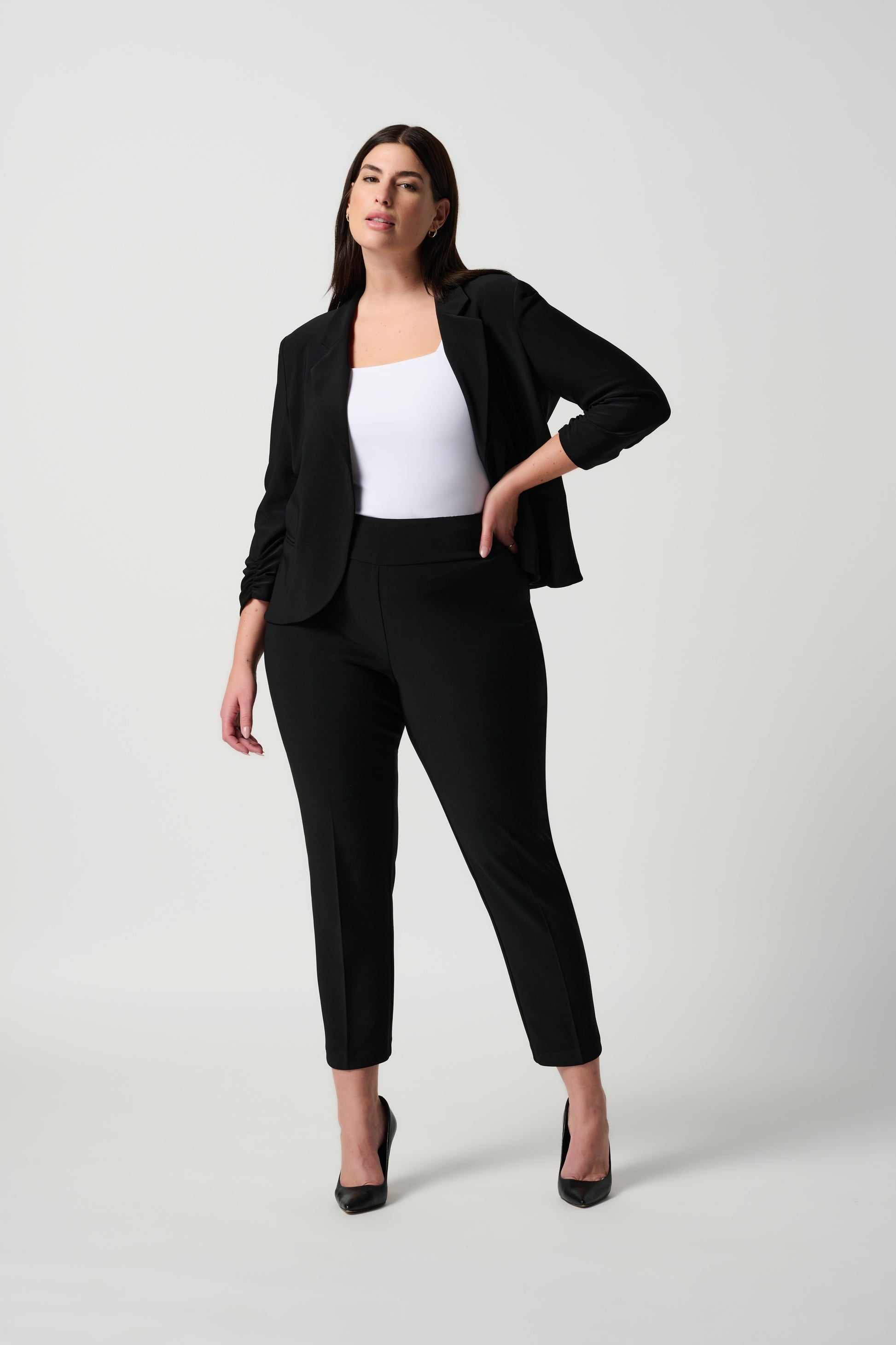 A person is donning Joseph Ribkoff's Classic Cropped Pant paired with black high-heeled shoes. The top portion of their outfit is a white shirt, and their arms rest by their sides. The background is plain and white.