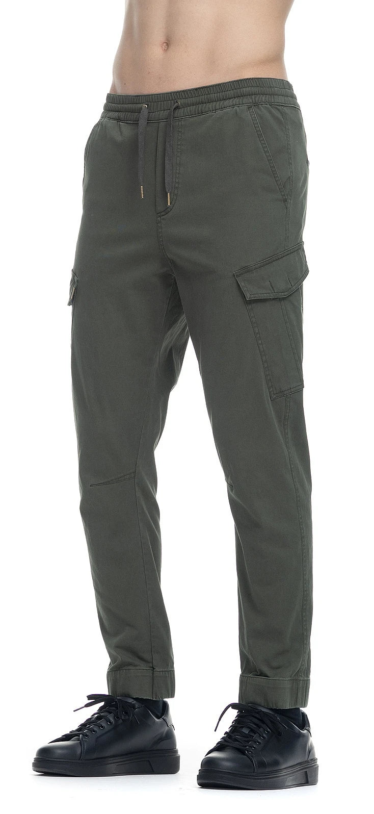 A person is wearing RAGWEAR's dark green ZEVL OLIVE trousers with multiple pockets and black sneakers. The vegan-certified pants feature an elastic waistband, and the hem is gathered at the ankles. The image is cropped to show the lower body only.