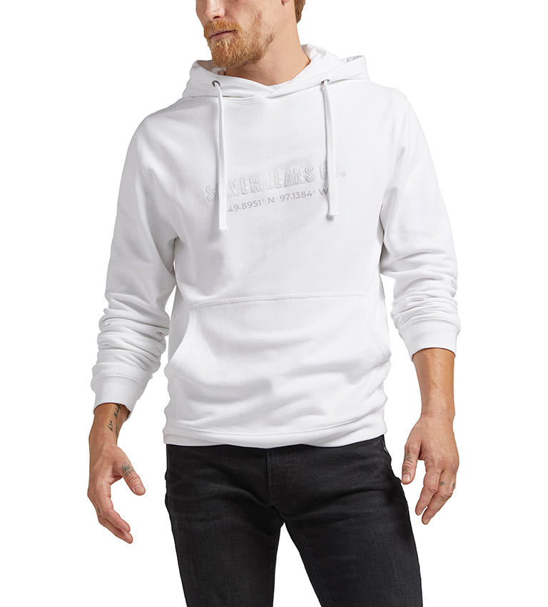 A person is wearing the Silver Jeans Co. Logo Hoodie - WHITE, a white French terry hoodie with drawstrings, a front pocket, and an embroidered logo featuring text and coordinates on the front. The 100% cotton hoodie pairs perfectly with their black jeans and beard. Only the upper body and part of the legs are visible.