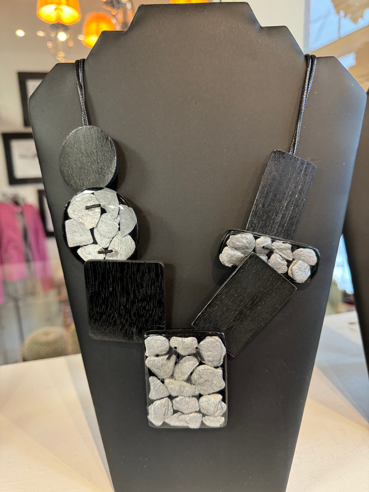 The Black Chunky Necklace by Posies Flowers & Fashion Inc. showcases an eclectic mix of rectangular and circular pendants, each embedded with clusters of small, textured grey stones. Presented as a striking statement piece, it is elegantly displayed on a matte black stand against a white background.