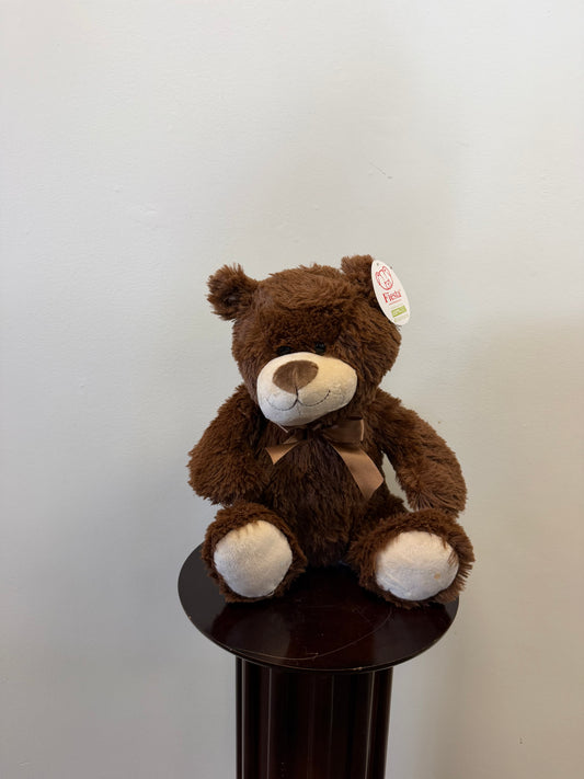 A Classic Teddy Bear - Brown 13 inch from Fiesta, featuring a white snout and paws with a ribbon and an ear tag, sits on a small round dark wooden table against a plain light-colored wall—making it an adorable choice for gift delivery.