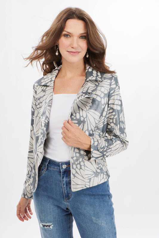 A woman with long brown hair is wearing a Joseph Ribkoff Printed Moto Jacket Style 231911, which features a butterfly-inspired motif, over a white top and blue jeans. She is smiling and looking at the camera while holding the jacket's lapel. The background is plain white.