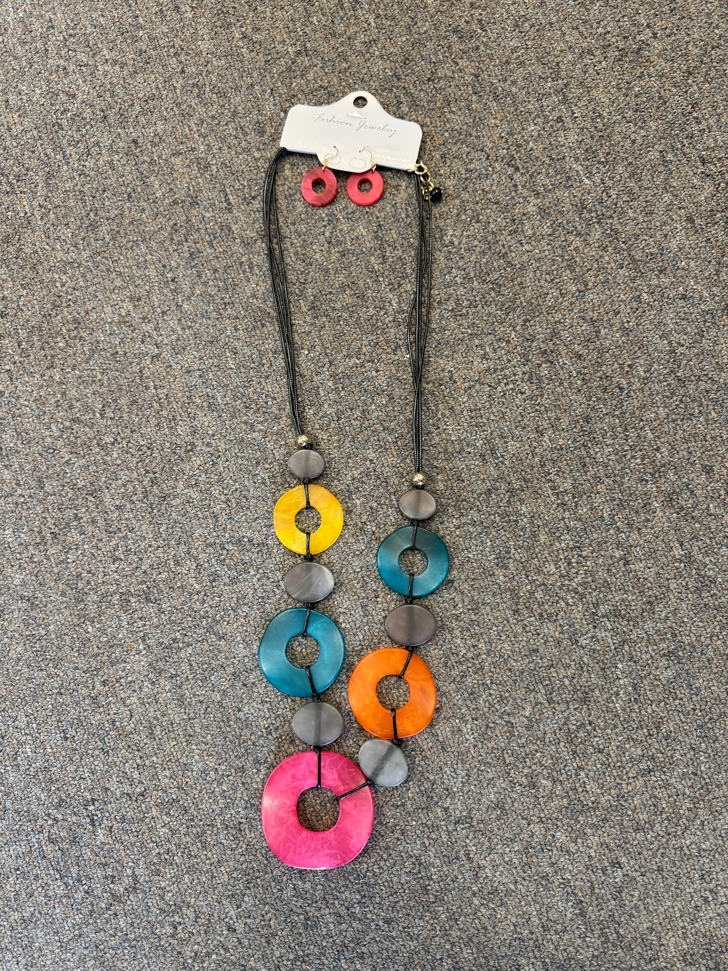 A colorful jewelry set from Posies Flowers & Fashion Inc., including the Rainbow Loops Necklace and Earrings, is displayed on a gray background. The necklace features large circular beads in pink, orange, yellow, and blue, interspersed with smaller gray beads. Matching pink circular earrings are attached to the packaging card of the necklace.