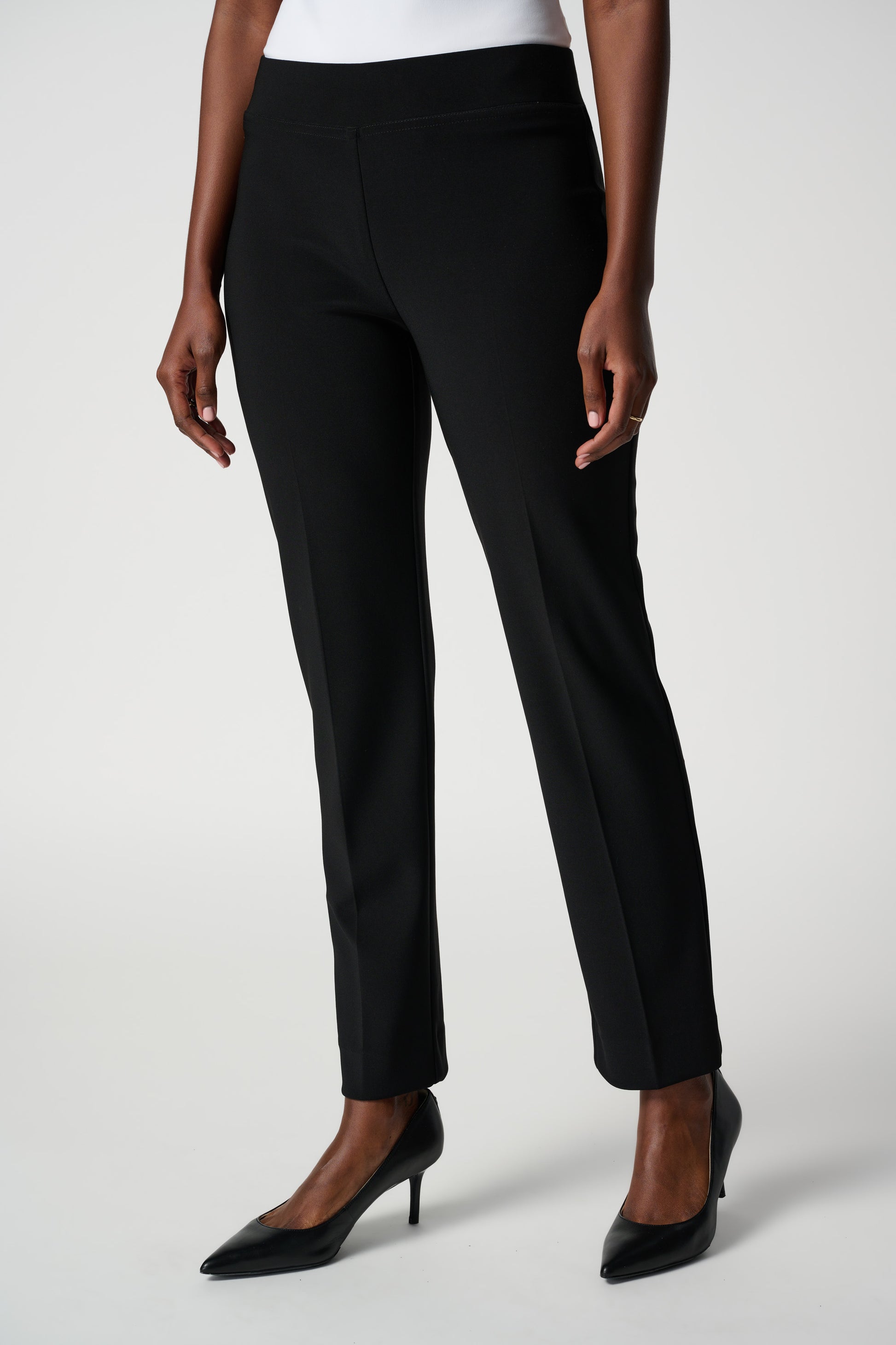 A person wearing the Classic Straight Pant in black by Joseph Ribkoff with slits at the ankles and black pointed heels, standing with their back to the camera. The person is also wearing a white top made of silky knit material. The background is plain white.