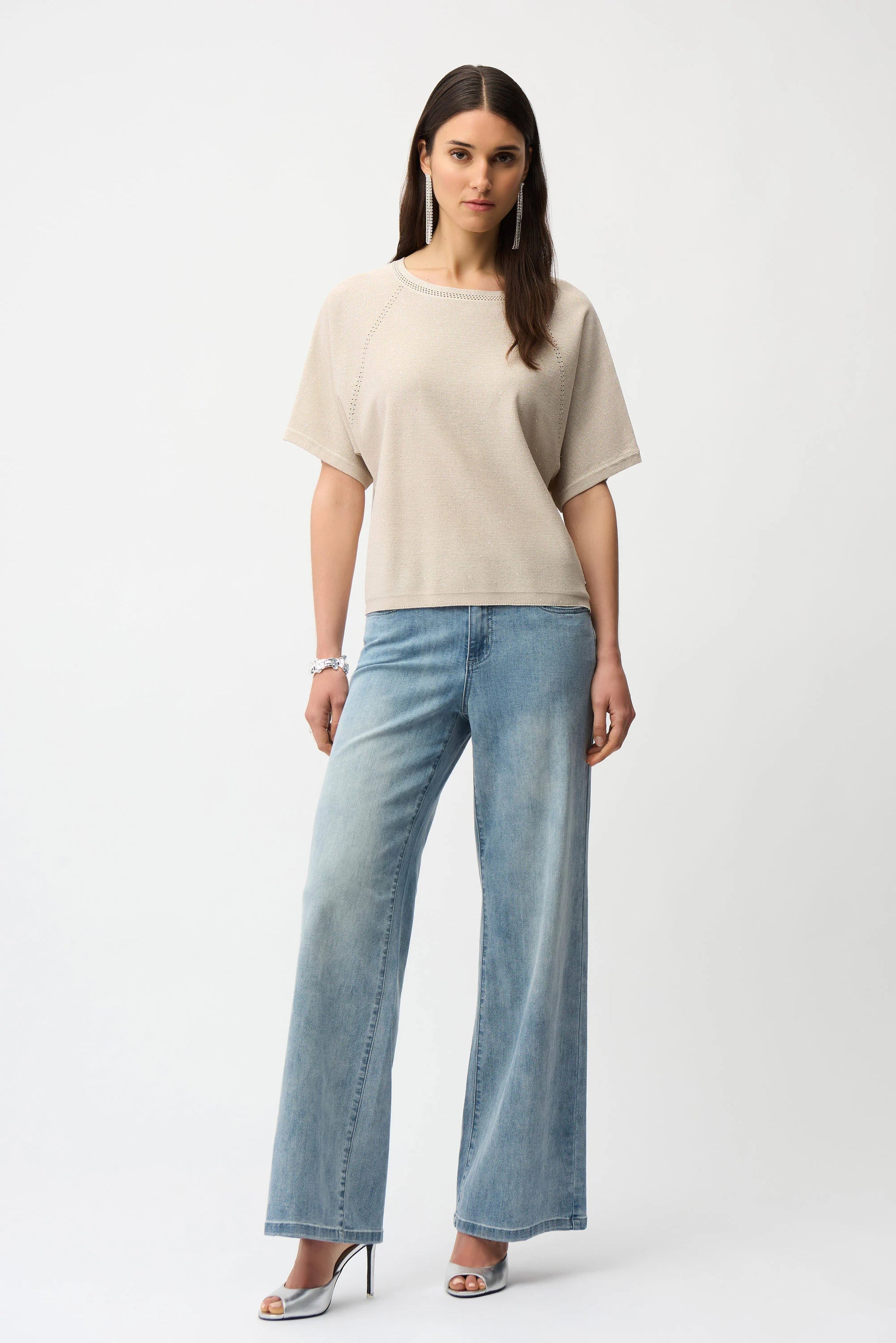 A woman with long, straight brown hair and a neutral expression wears the Joseph Ribkoff Sequins Yarn Short Sleeve Sweater 261917 against a plain white background. She pairs it with light blue wide-leg jeans, silver heels, and hoop earrings.