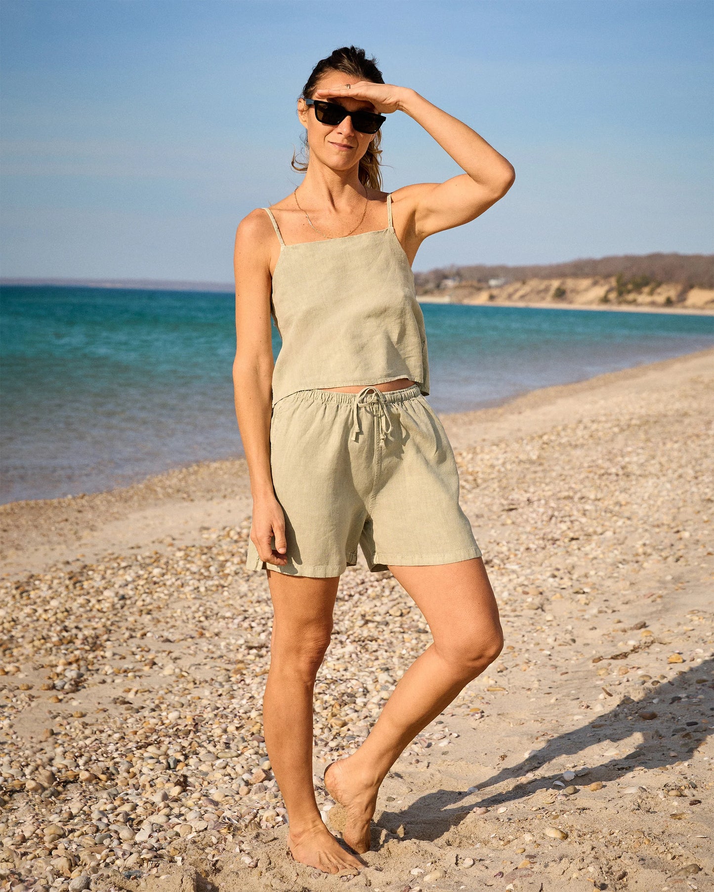A woman in the Alex Crane Women's Penny Cami / Seagrass stands barefoot on a pebbly beach, shading her eyes with one hand and wearing sunglasses, with the blue sea and distant shoreline behind her.